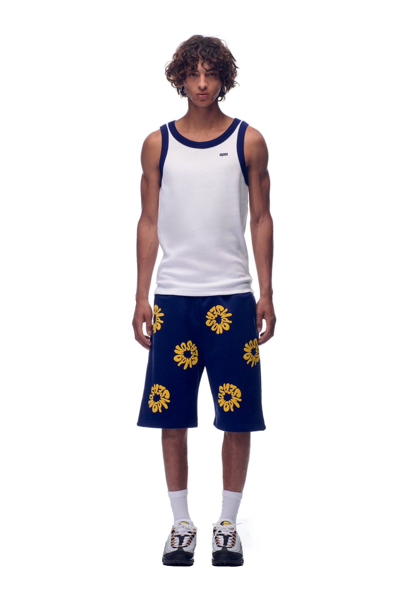 A young man with curly hair models the 6PM NH TANKTOP WHITE/NAVY, paired with blue shorts featuring bold yellow floral designs, white socks, and black sneakers, standing against a plain white background.