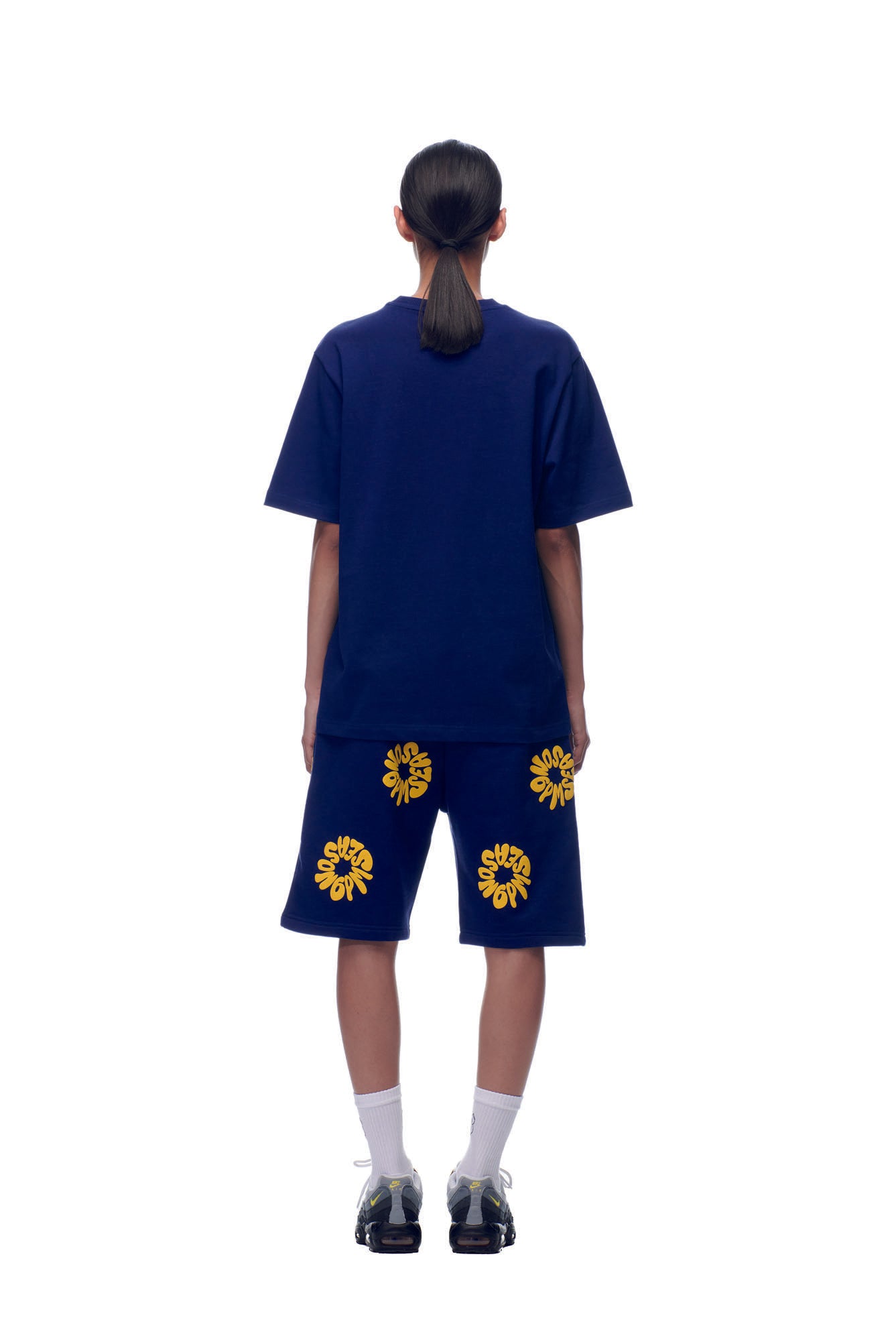 A person stands facing away on a white background, wearing the 6PM T-SHIRT FLOWER NAVY with matching shorts featuring yellow sun graphics, paired with white socks and black sneakers.