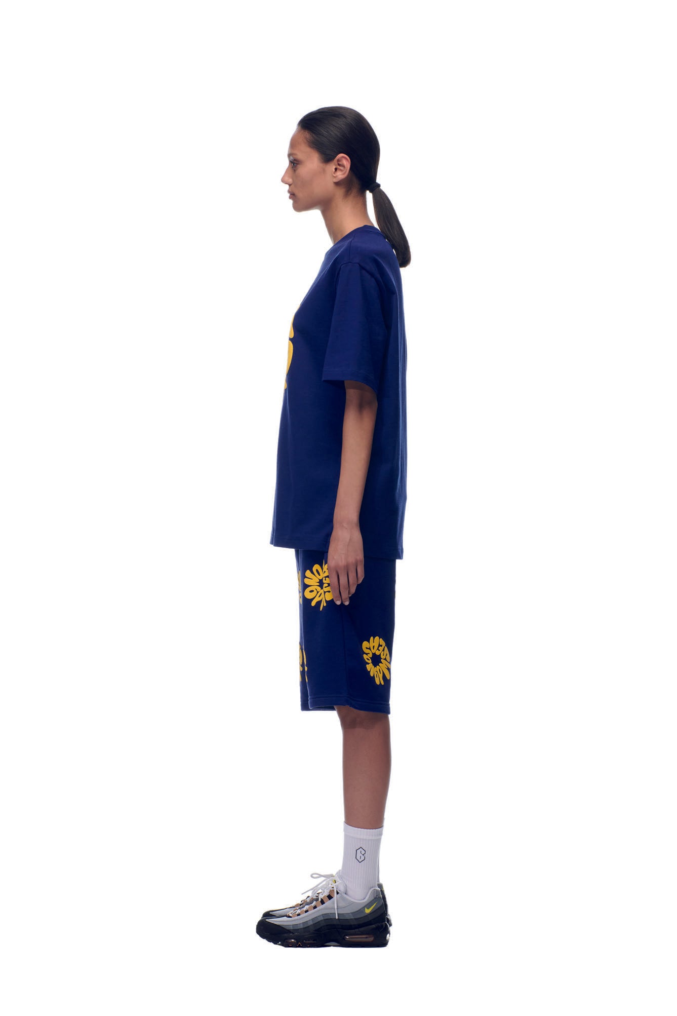 A woman in profile stands against a white background, wearing the 6PM T-SHIRT FLOWER NAVY and matching shorts with yellow flower prints, paired with white socks, black sneakers, and her dark hair in a low ponytail.