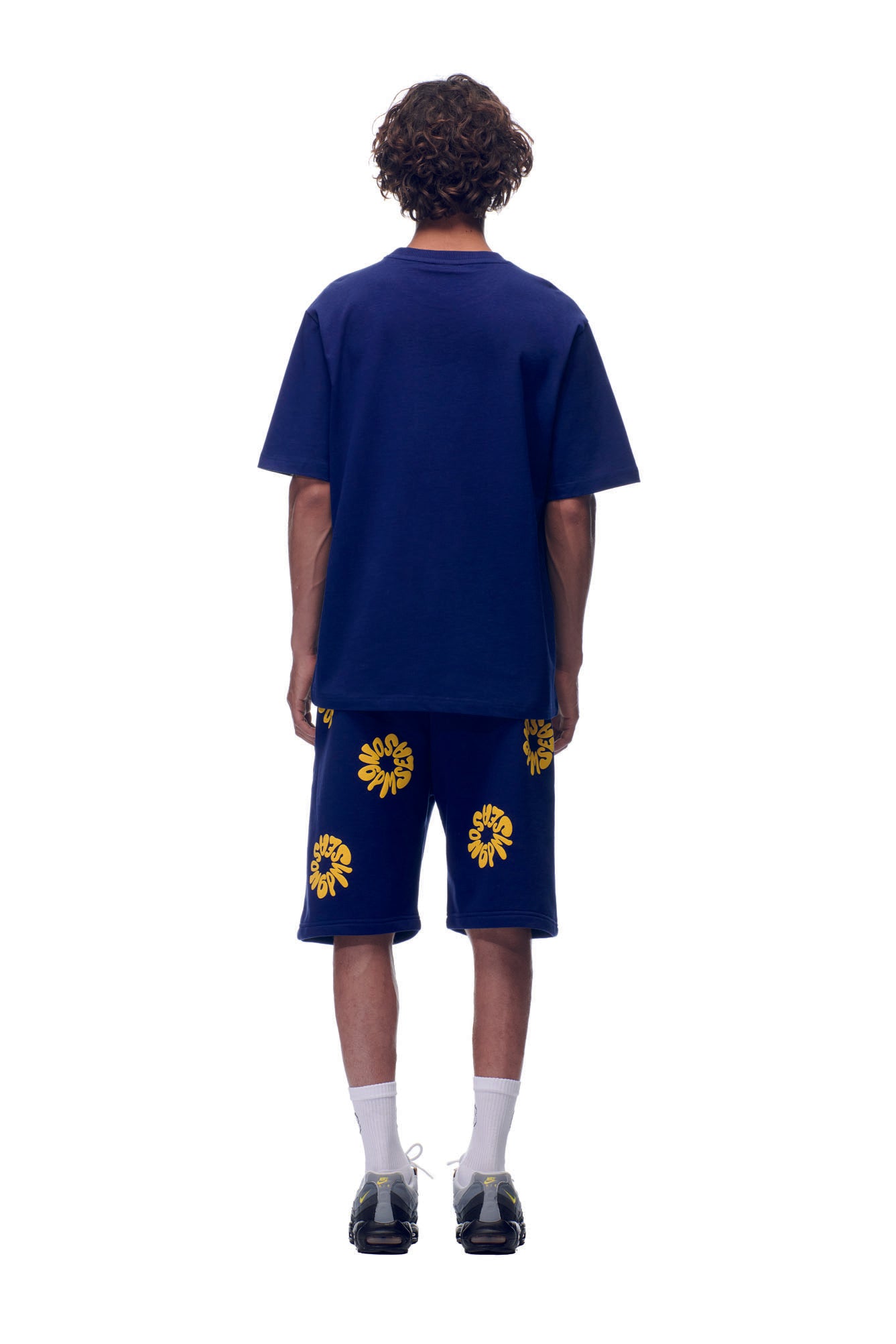 A person with curly hair stands against a white background, wearing the 6PM T-SHIRT FLOWER NAVY and matching shorts. The navy outfit features yellow flower patterns, paired with white socks and black sneakers.