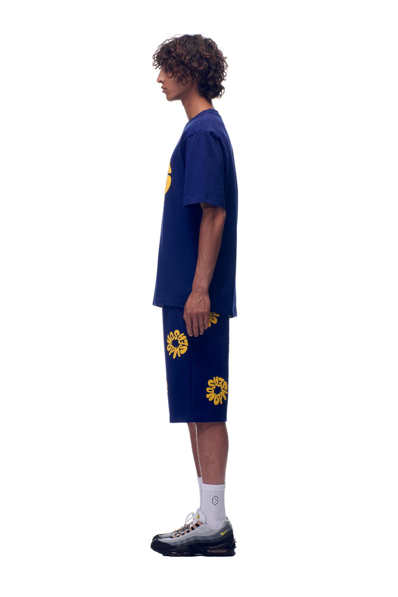 A young man stands in profile against a white background, wearing the 6PM T-SHIRT FLOWER NAVY with matching shorts featuring bold yellow graphics, paired with white socks and black sneakers.