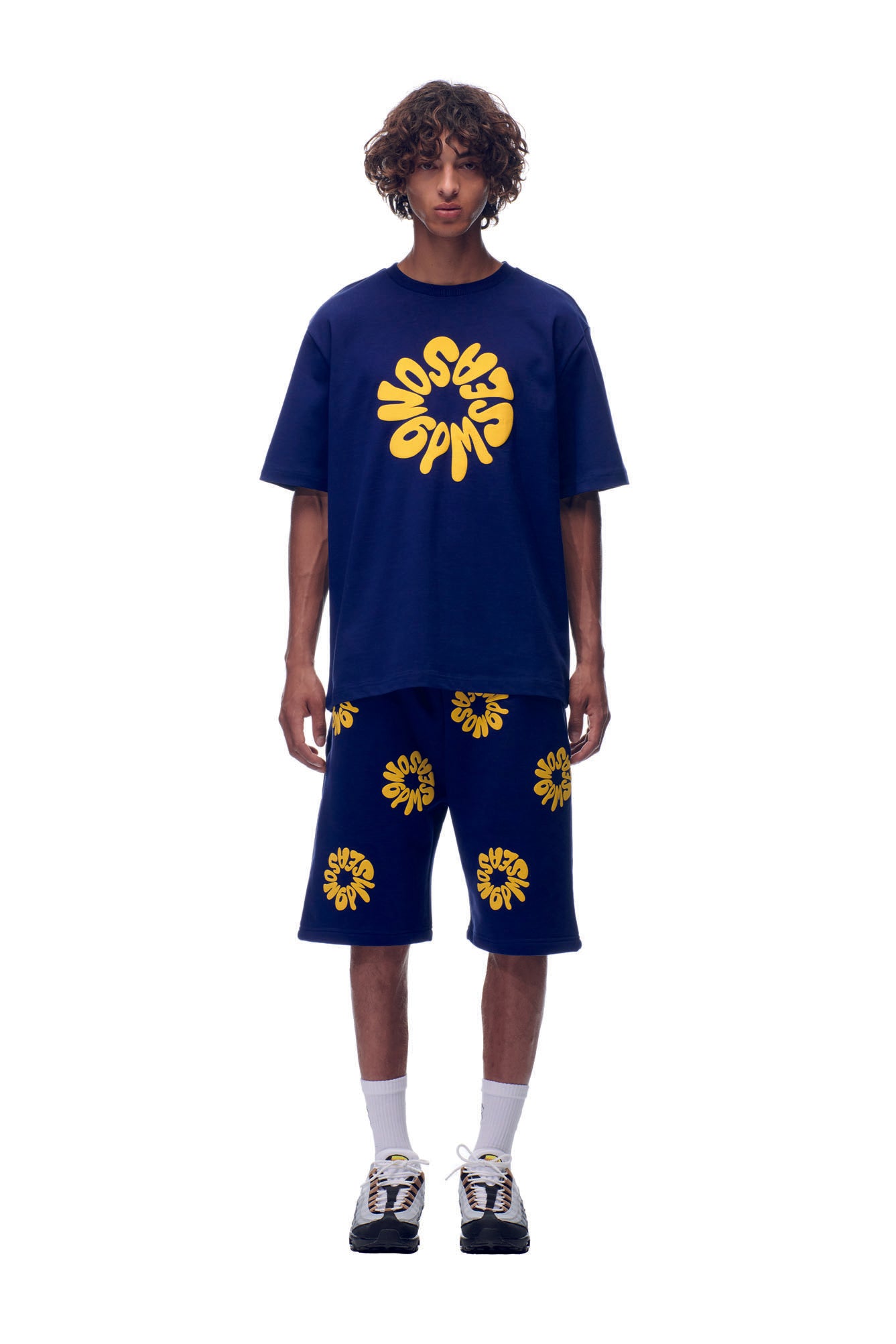 A person with curly hair stands against a plain white background, wearing the 6PM T-SHIRT FLOWER NAVY oversized tee and matching shorts featuring yellow abstract graphics, paired with white socks and sneakers.