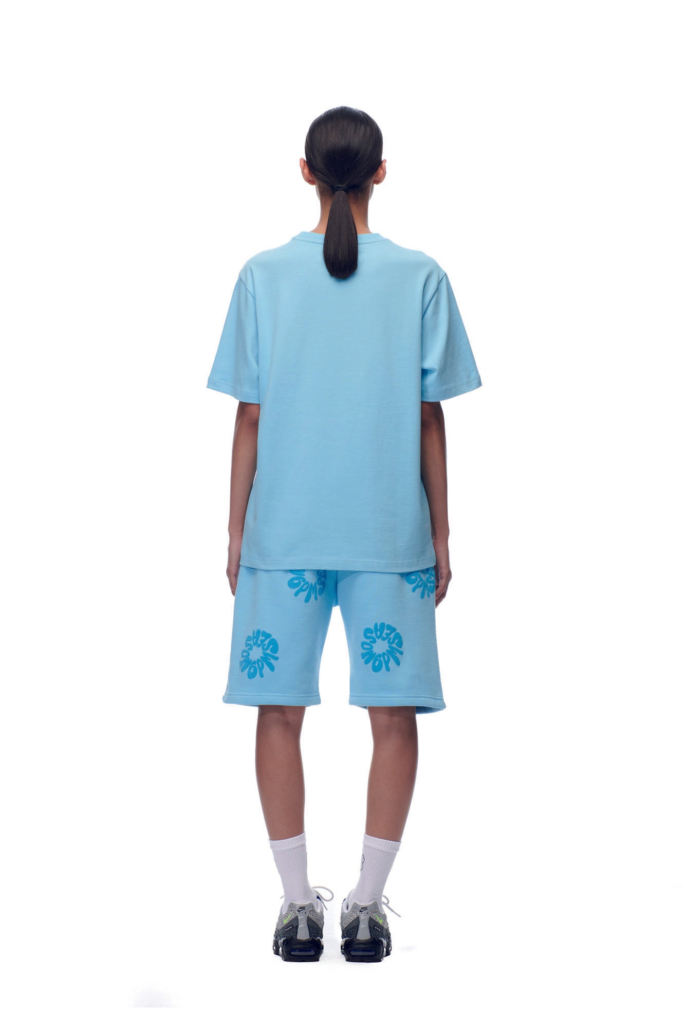 A person with a low ponytail, facing away, wears the 6PM SCAMMER T-SHIRT BABY BLUE paired with matching floral blue shorts, white socks, and black sneakers against a white background.