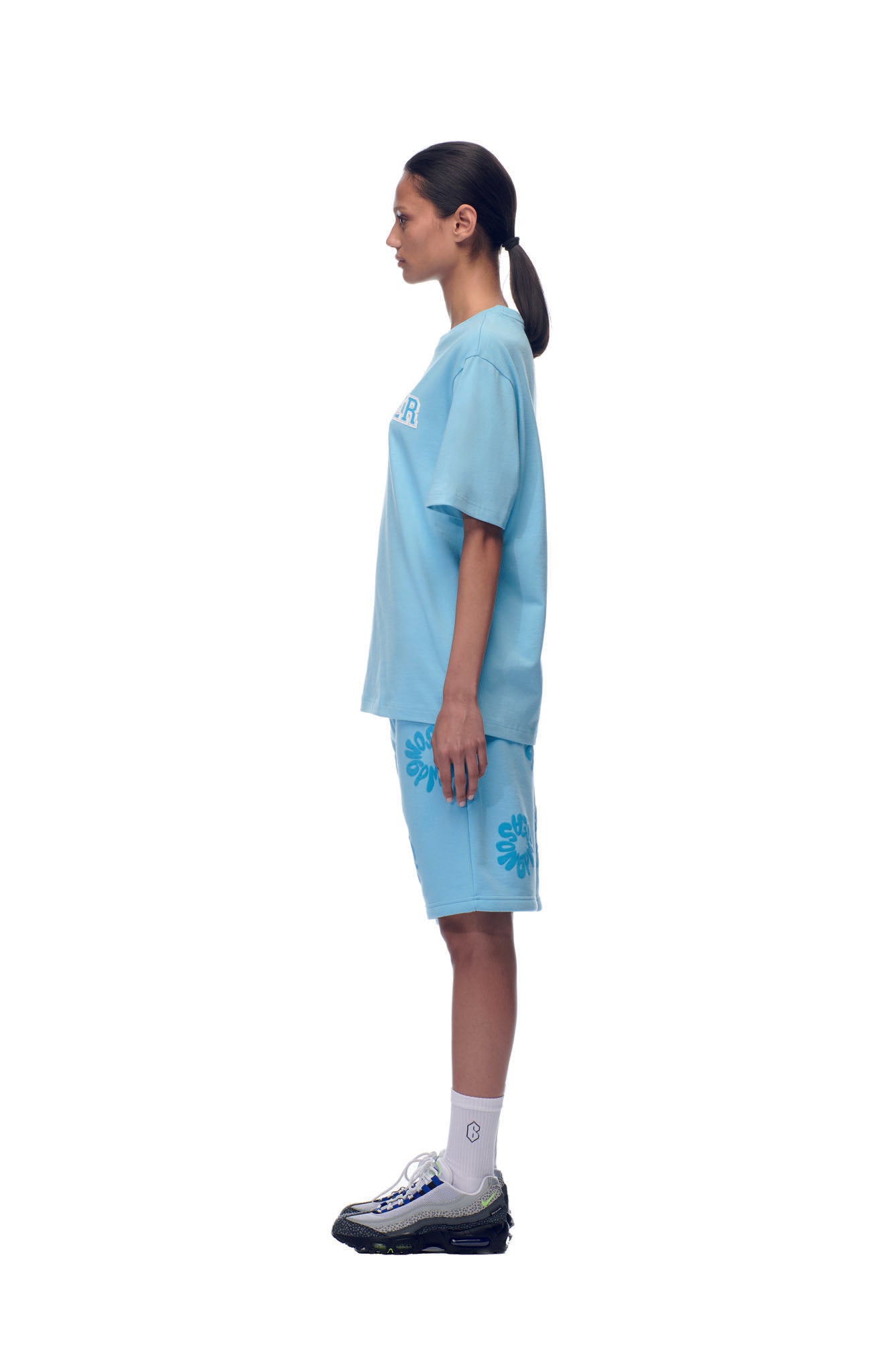 A person with dark hair in a low ponytail stands in profile, wearing the 6PM SCAMMER T-SHIRT BABY BLUE, matching blue floral shorts, white socks, and sporty sneakers against a white background.