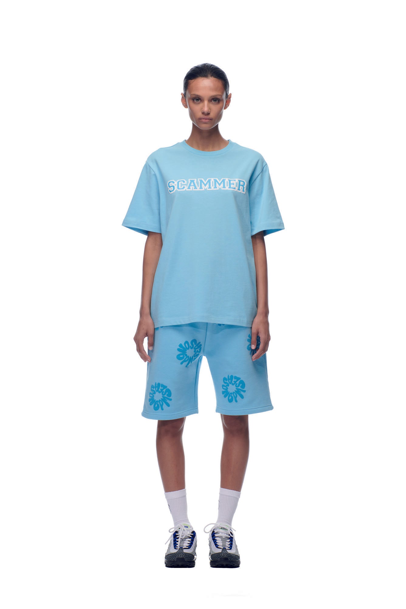 A person faces forward wearing the 6PM SCAMMER T-SHIRT BABY BLUE, paired with matching blue floral shorts, white socks, and blue athletic shoes against a white background.