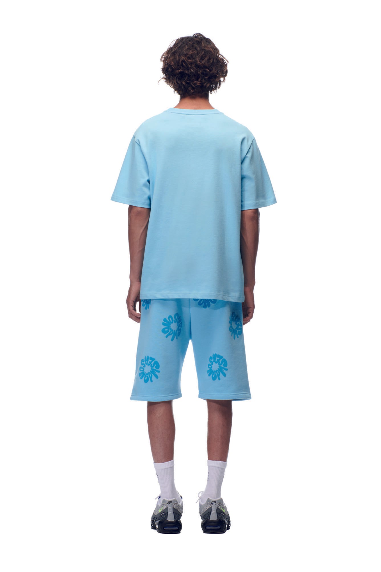 A person with curly hair faces away, wearing the 6PM SCAMMER T-SHIRT in baby blue, matching blue leaf print shorts, white socks, and black sneakers against a plain white background.