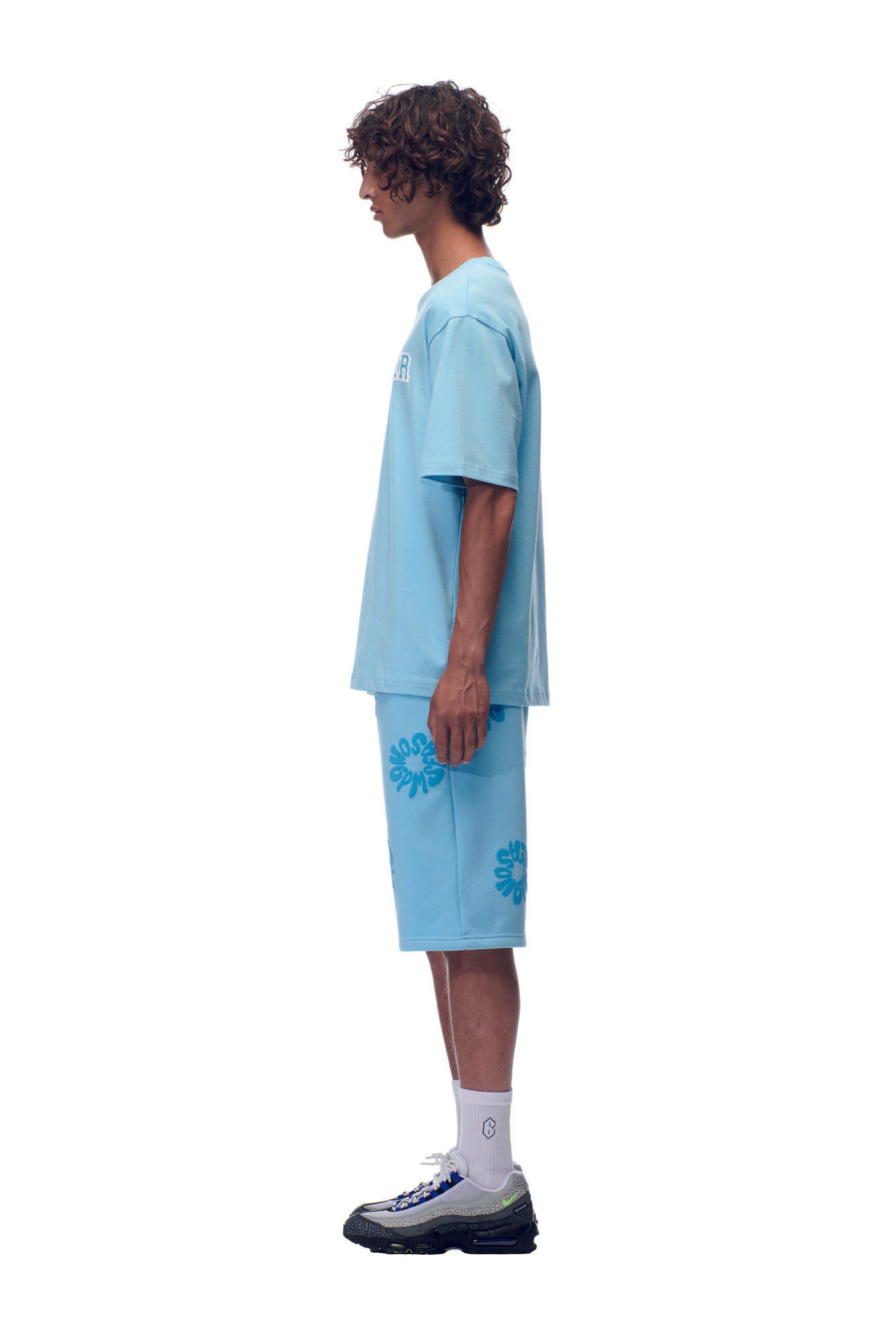 A young man with curly hair stands in profile, wearing the 6PM SCAMMER T-SHIRT BABY BLUE and matching shorts with blue graphics, white socks, and black sneakers, set against a plain white background.