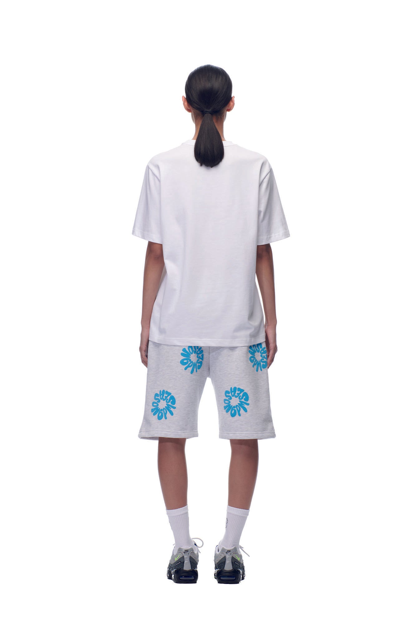 A person with long dark hair in a low ponytail faces away, wearing the 6PM NH PLAY LOGO T-SHIRT WHITE, gray shorts with blue floral designs, white socks, and black sneakers against a white background.