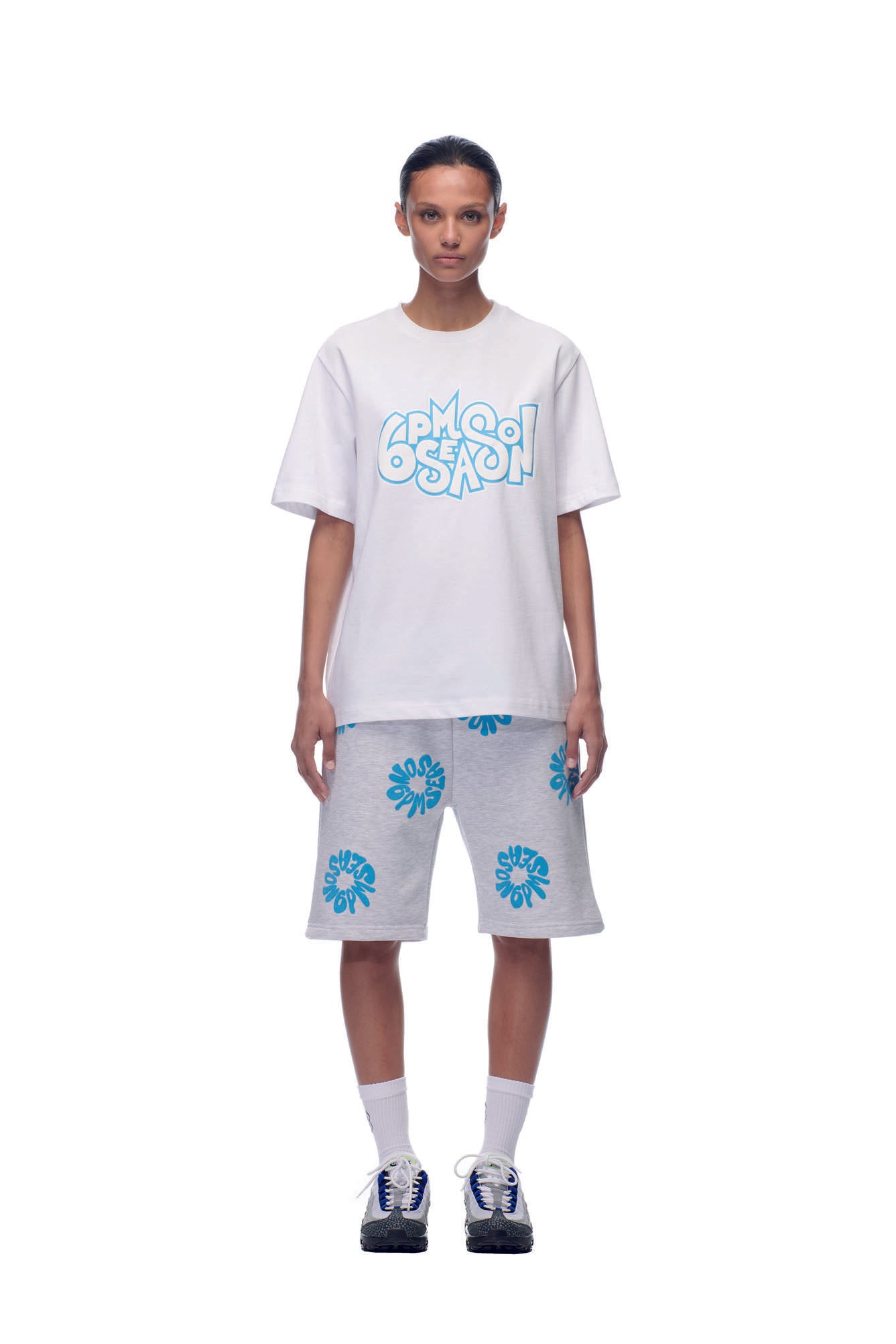 A person stands against a white background wearing the 6PM NH PLAY LOGO T-SHIRT WHITE, paired with gray knee-length shorts featuring blue designs, as well as white socks and sneakers.