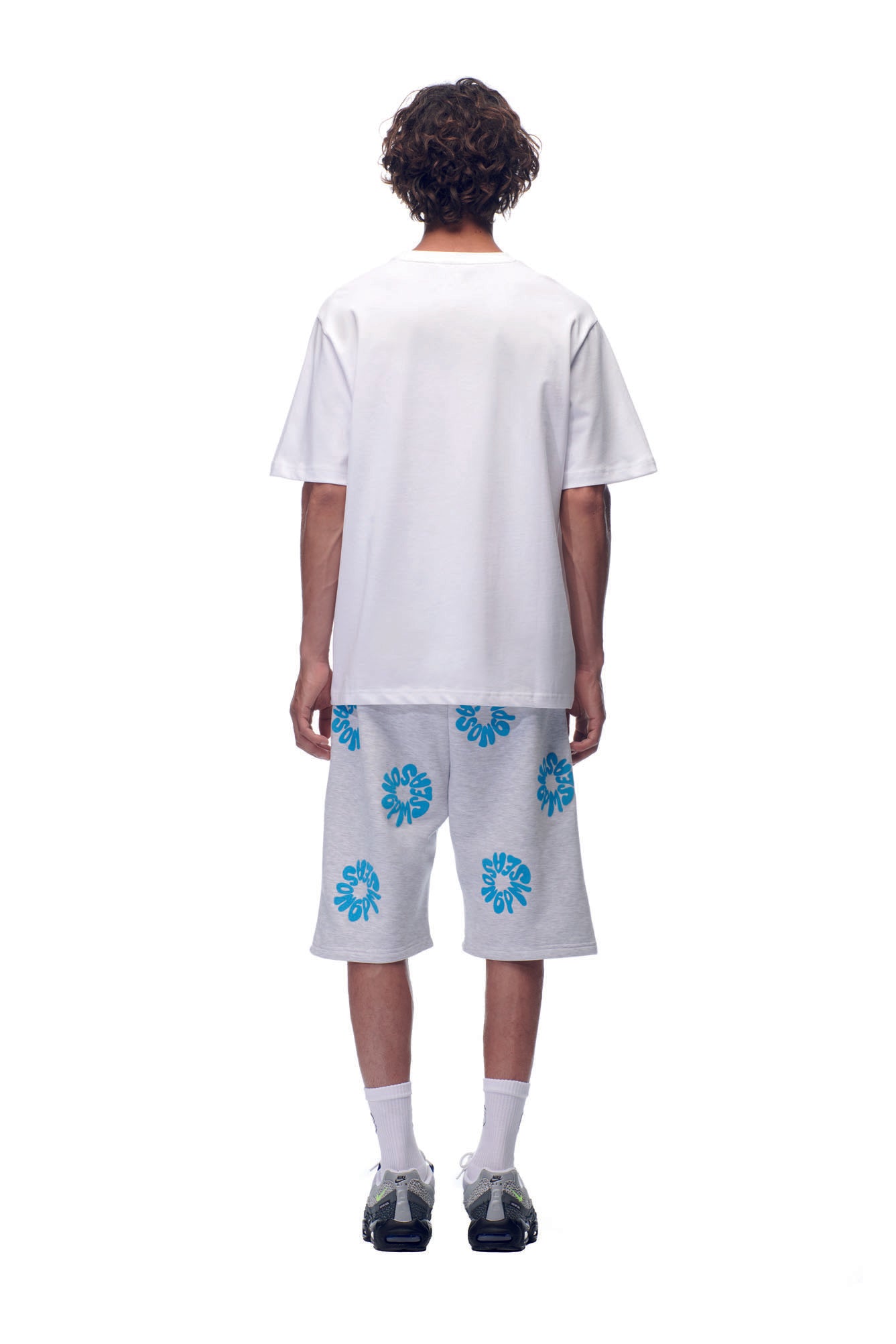 A person faces away in a 6PM NH PLAY LOGO T-SHIRT WHITE, paired with light gray shorts featuring blue sunburst patterns, white socks, and black sneakers, against a plain white background.