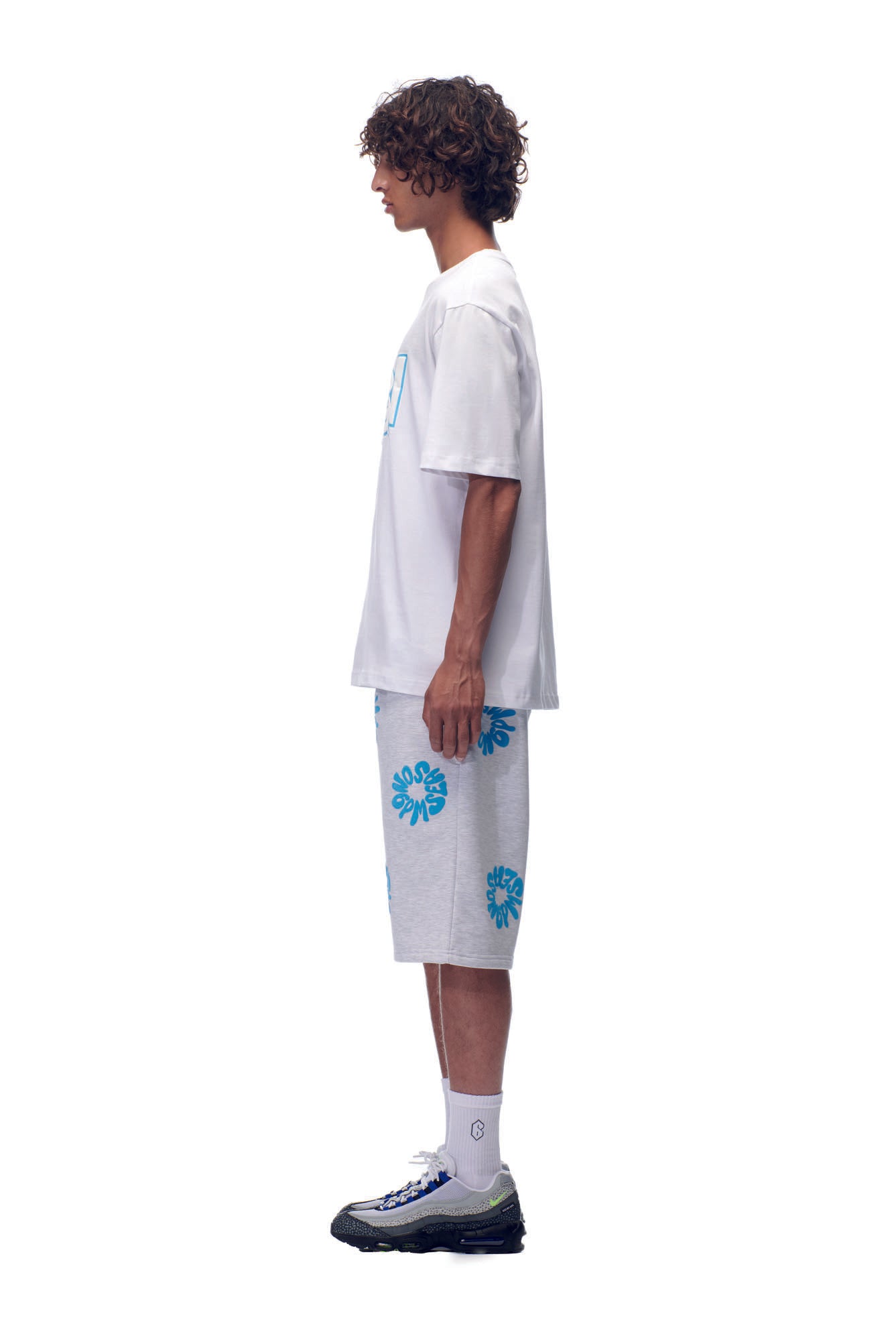 A person stands in profile against a white background, wearing the NH PLAY LOGO T-SHIRT WHITE by 6PM, light gray shorts with blue floral patterns, white socks, and gray athletic shoes with black soles.
