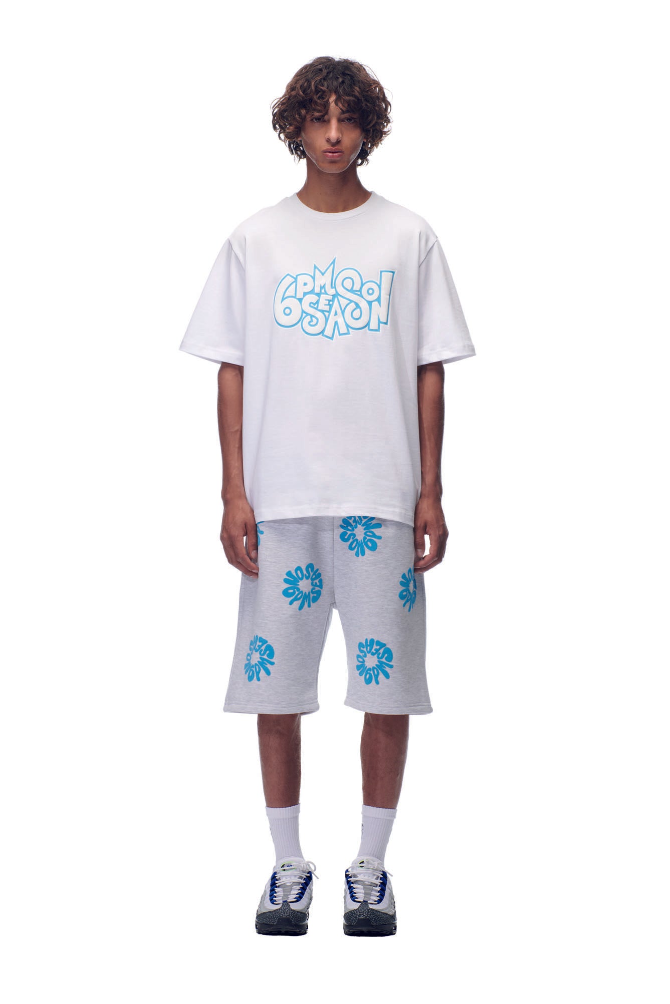 A young man faces forward against a white background, wearing the 6PM NH PLAY LOGO T-SHIRT in white, gray knee-length shorts with blue designs, white socks, and black-and-white sneakers.