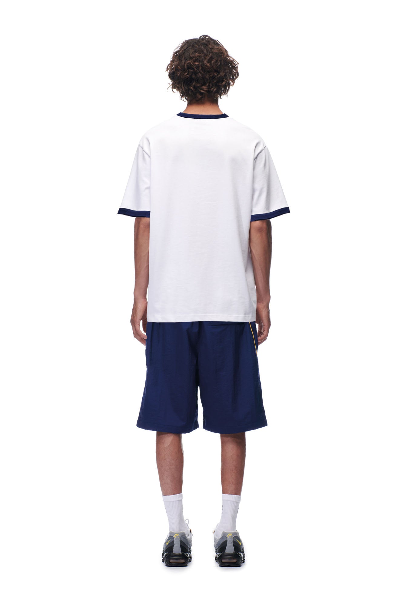 A person stands facing away, wearing the 6PM SCAMMER T-SHIRT WHITE with navy trim, navy shorts, white socks, and black sneakers against a plain white background.