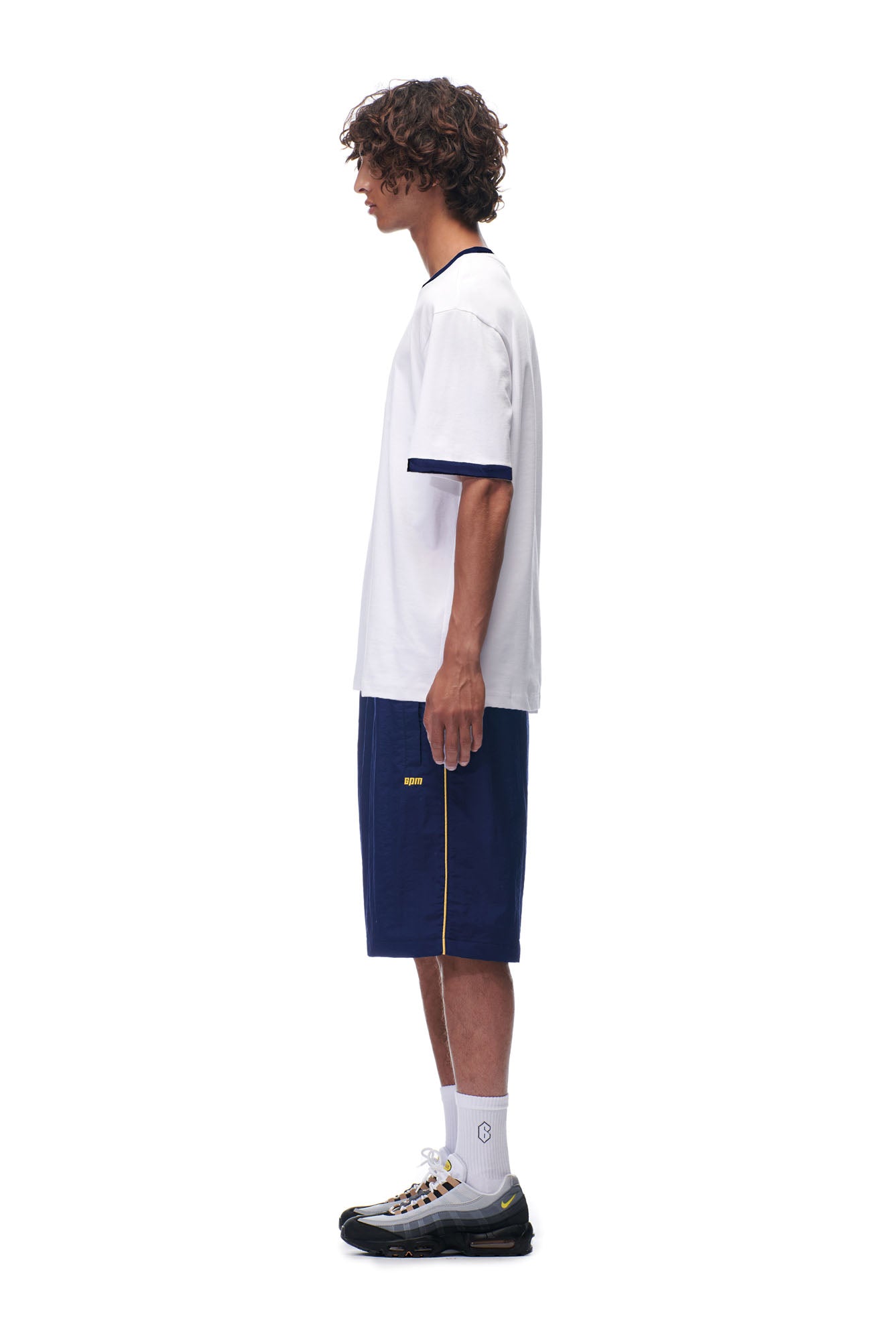 A young man with curly hair stands in profile against a white background, wearing the 6PM SCAMMER T-SHIRT WHITE, navy blue shorts with yellow trim, white socks, and black sneakers.