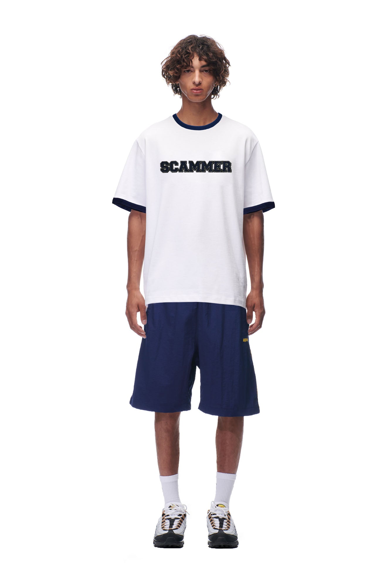 A young man with curly hair wears the 6PM SCAMMER T-SHIRT WHITE, paired with navy blue shorts, white socks, and sneakers, against a plain white background.