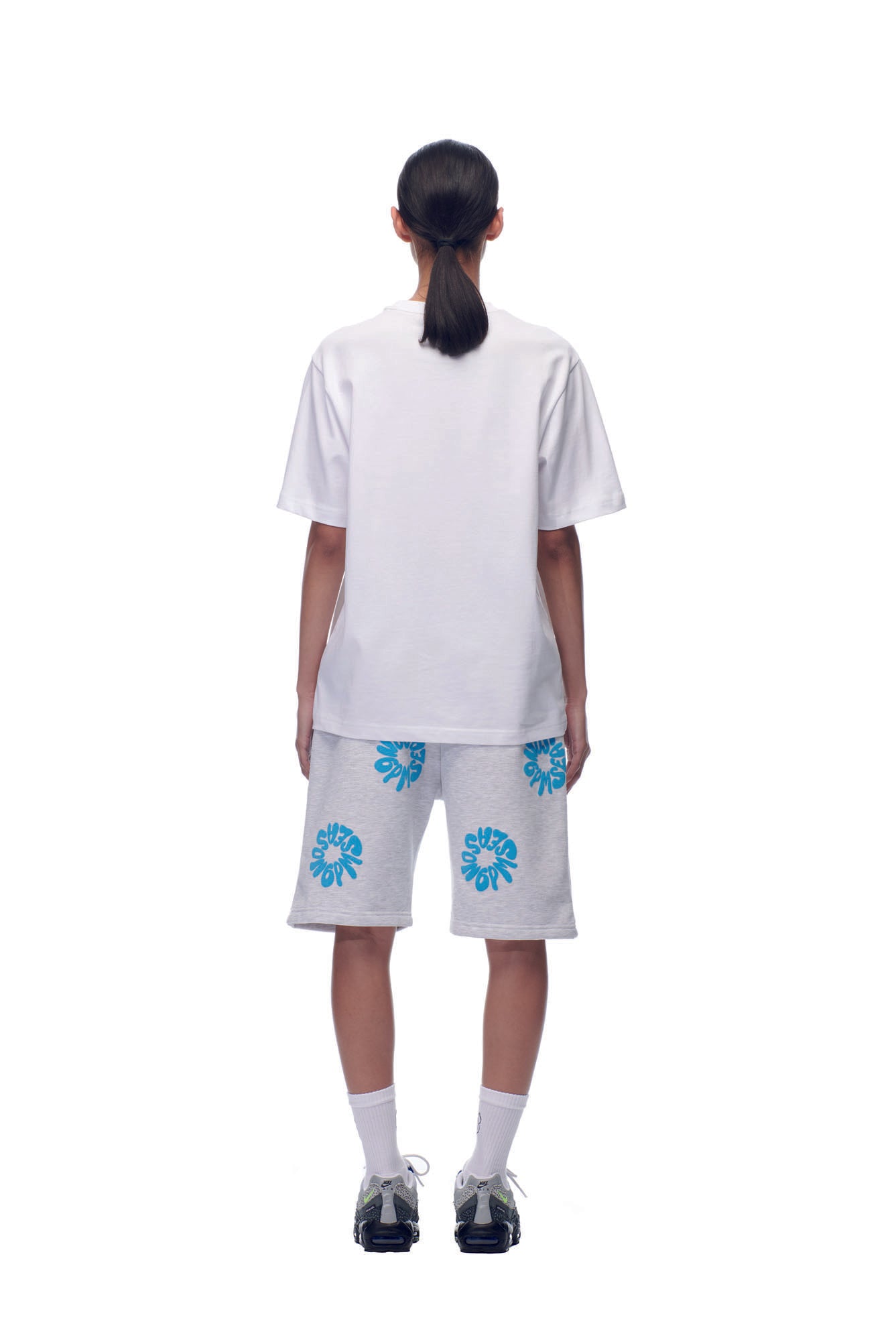A person stands with their back to the camera, wearing a 6PM SELL HOPE T-SHIRT WHITE, white shorts with blue floral patterns, white socks, and black sneakers against a plain white background.