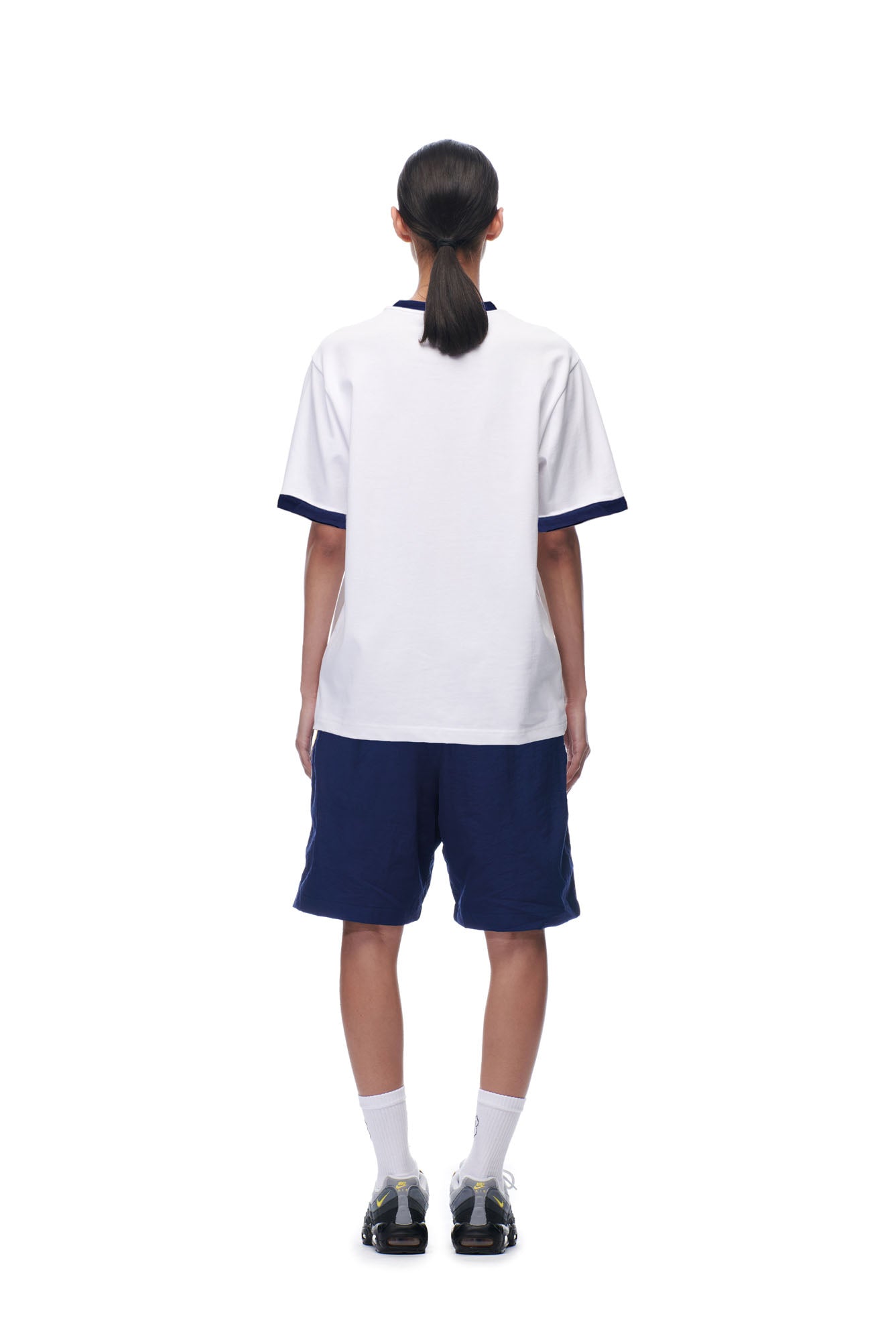 A person stands with their back to the camera, wearing the 6PM SCAMMER T-SHIRT WHITE, navy blue shorts, white socks, black sneakers, and a low ponytail. The background is white.
