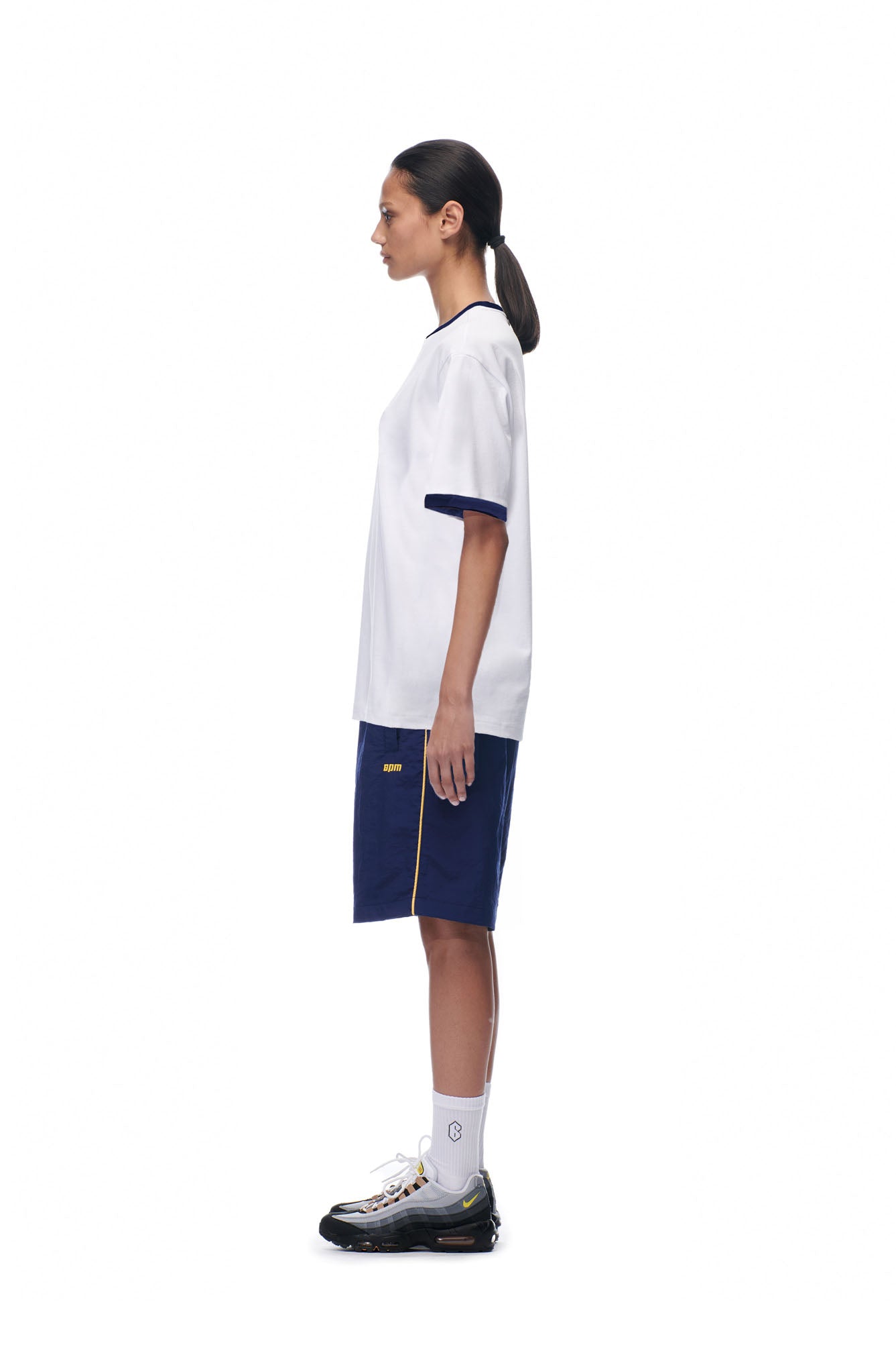 A person stands in profile against a white background, wearing the 6PM SCAMMER T-SHIRT WHITE, navy athletic shorts with a yellow stripe, white socks, and black sneakers with white soles.