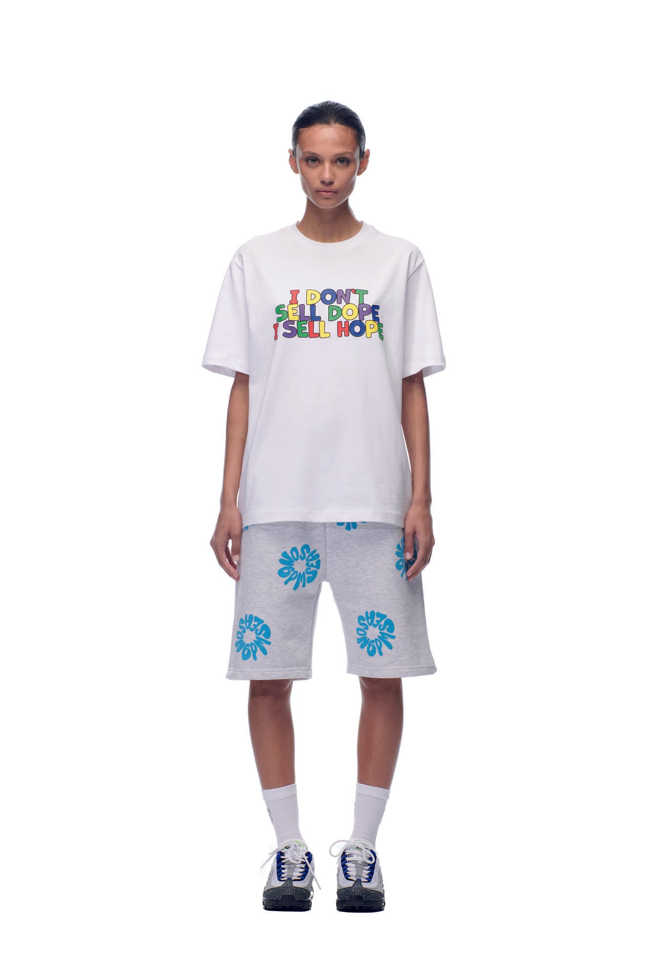 A woman faces forward wearing the 6PM SELL HOPE T-SHIRT WHITE with colorful "I DONT SELL DOPE I SELL HOPE" text, paired with light gray shorts featuring blue flowers, white socks, and black sneakers.