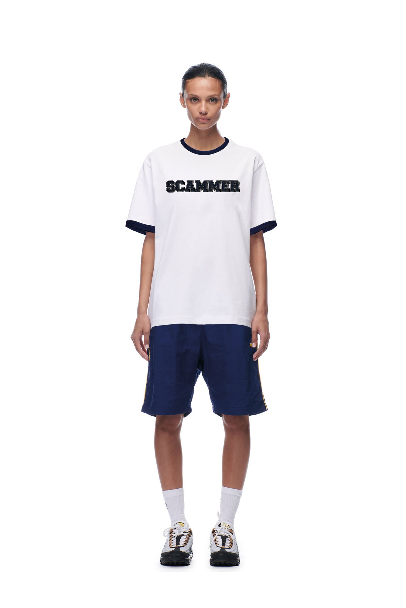 A person faces forward against a plain white background wearing the 6PM SCAMMER T-SHIRT WHITE, paired with navy blue shorts, white socks, and sneakers.