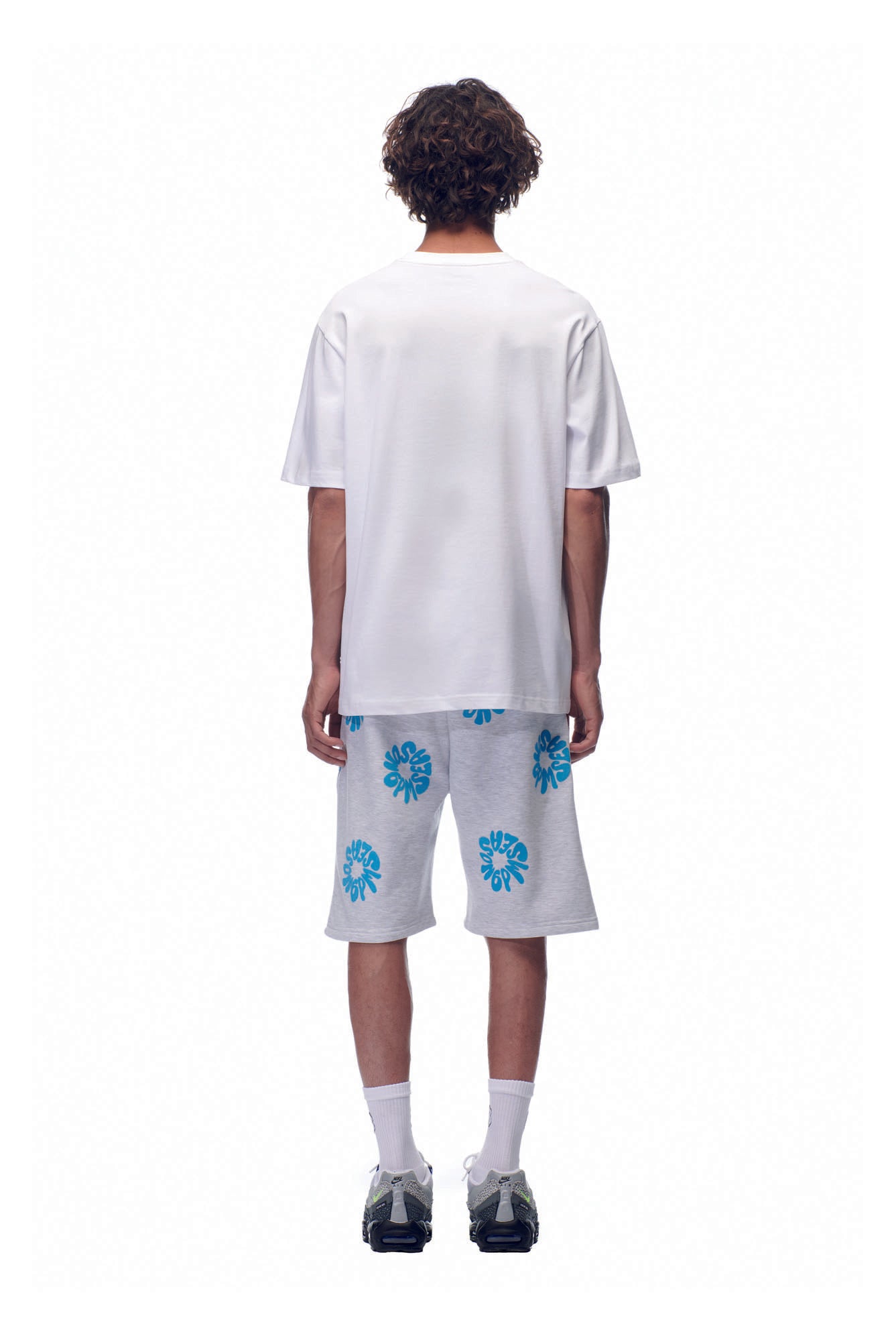 A person stands with their back to the camera, wearing the 6PM SELL HOPE T-SHIRT WHITE, paired with white knee-length shorts featuring blue floral patterns, white socks, and dark sneakers against a plain white background.