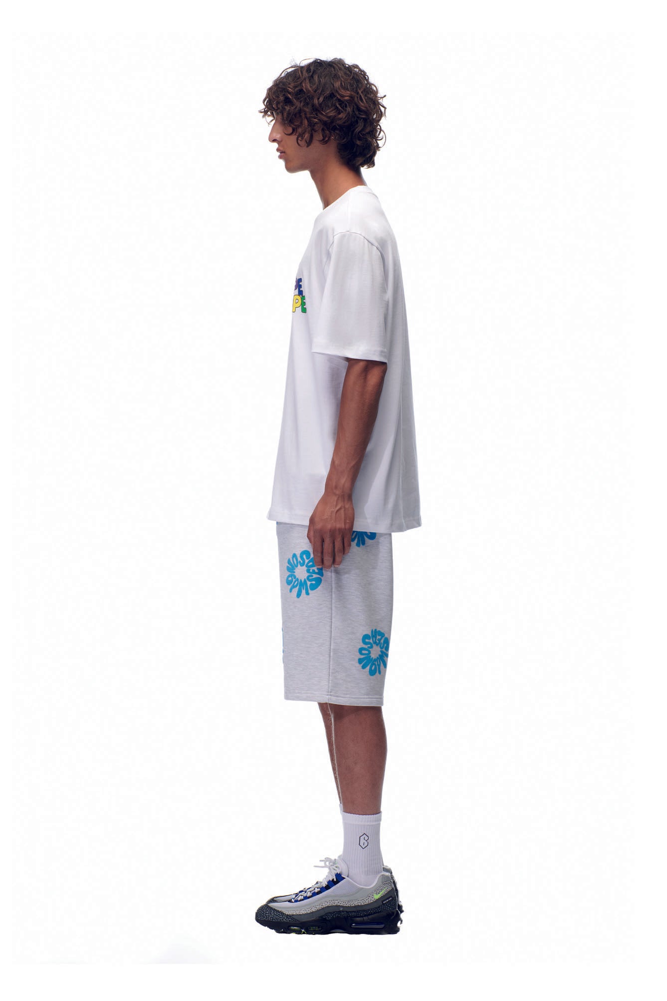 A young man with curly hair stands in profile, wearing the 6PM SELL HOPE T-SHIRT WHITE, light gray knee-length shorts with blue flower designs, white socks, and gray sneakers against a plain white background.