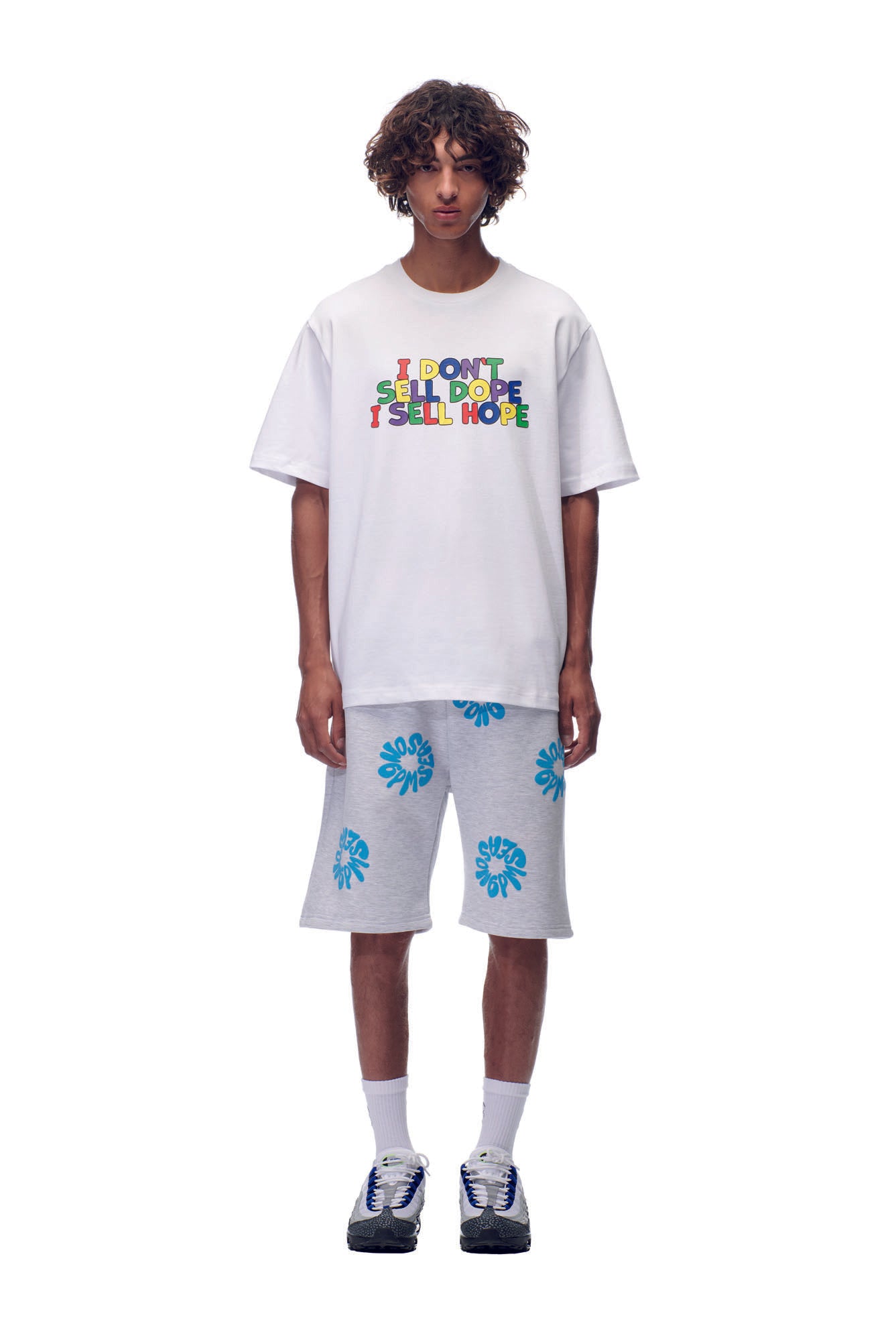 A person wears the 6PM SELL HOPE T-SHIRT WHITE with colorful “I DON’T SELL DOPE I SELL HOPE” text, paired with light gray shorts featuring blue leaf prints, plus white socks and sneakers, against a white background.