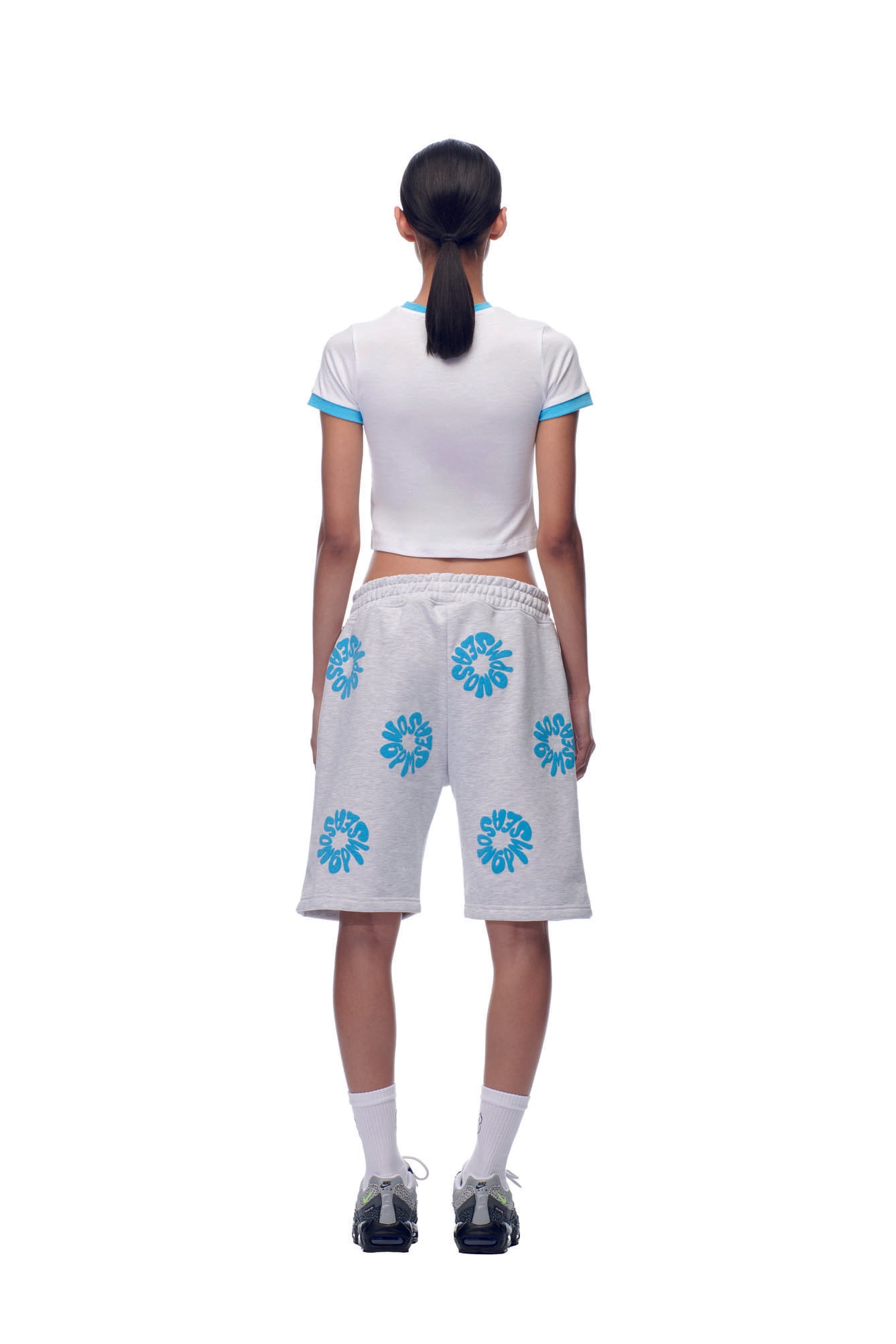 A person faces away, wearing the 6PM NH LOGO BABY TEE WHITE with white knee-length shorts featuring blue floral patterns, white socks, and black sneakers, standing against a plain white background.