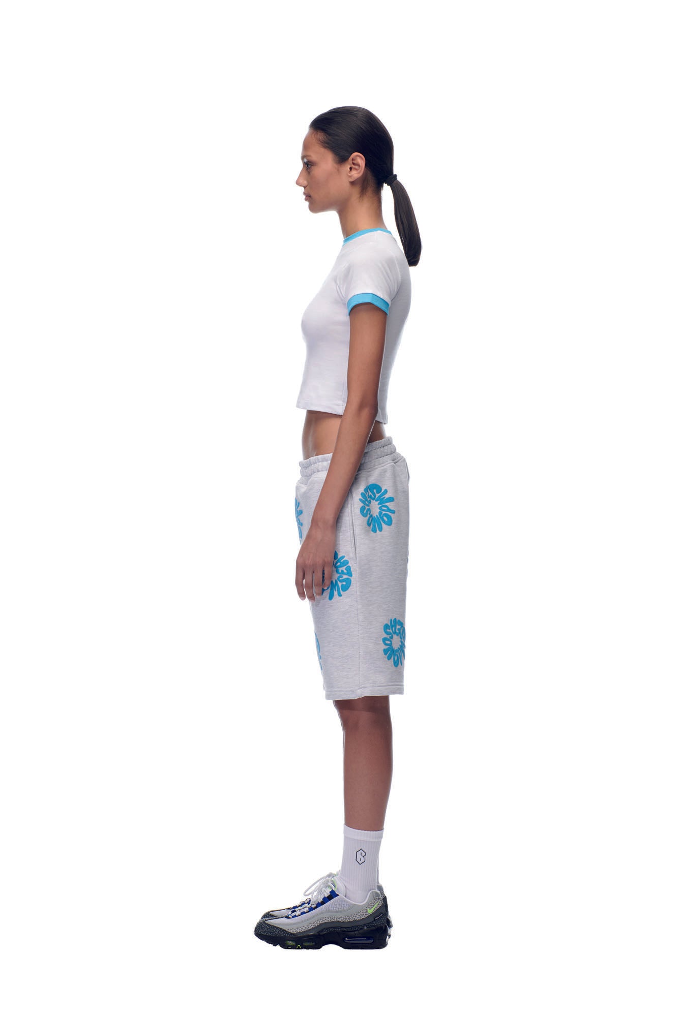 A woman stands in profile against a white background, wearing the 6PM NH LOGO BABY TEE WHITE, grey knee-length shorts with blue floral prints, white socks, sneakers, and her dark hair tied back in a low ponytail.