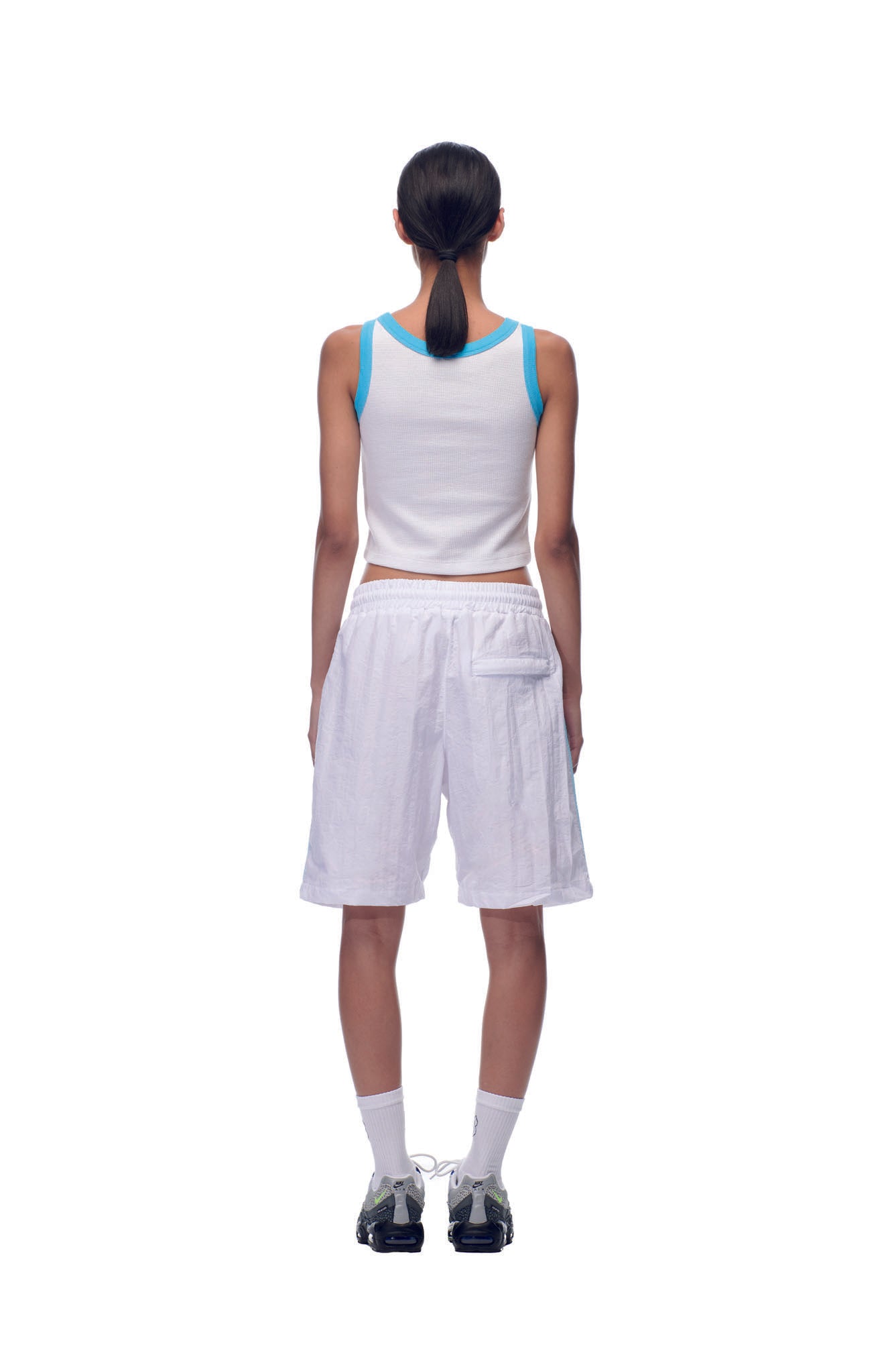 A person stands facing away, wearing NH NYLON SHORTS WHITE by 6PM with a white sleeveless top, blue trim, white socks, and black sneakers. Their hair is tied in a low ponytail against a plain white background.