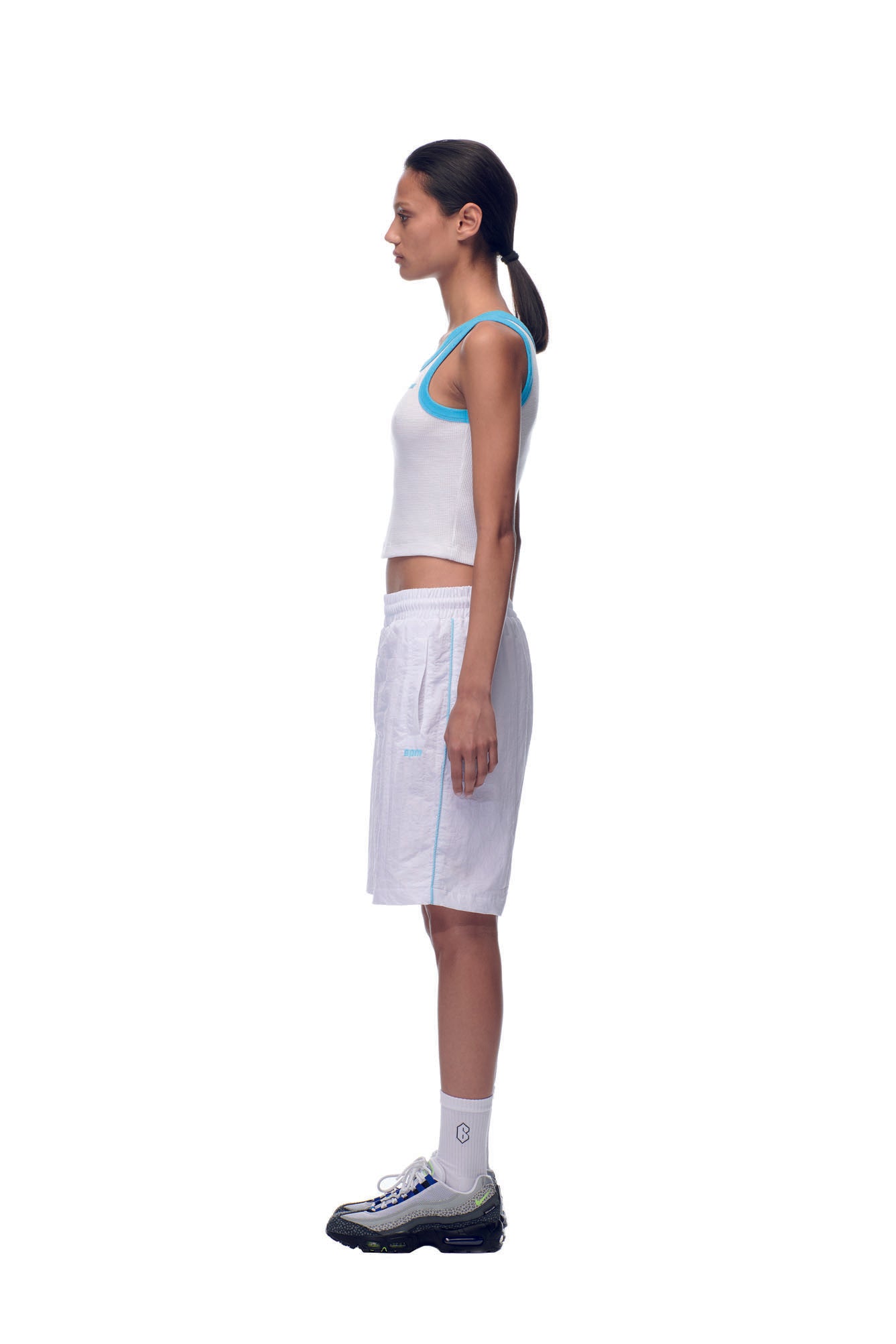 A woman stands in profile against a white background, wearing 6PM's NH Nylon Shorts in white along with a white tank top, blue trim, crew socks, black and white sneakers, and her hair pulled back in a low ponytail.