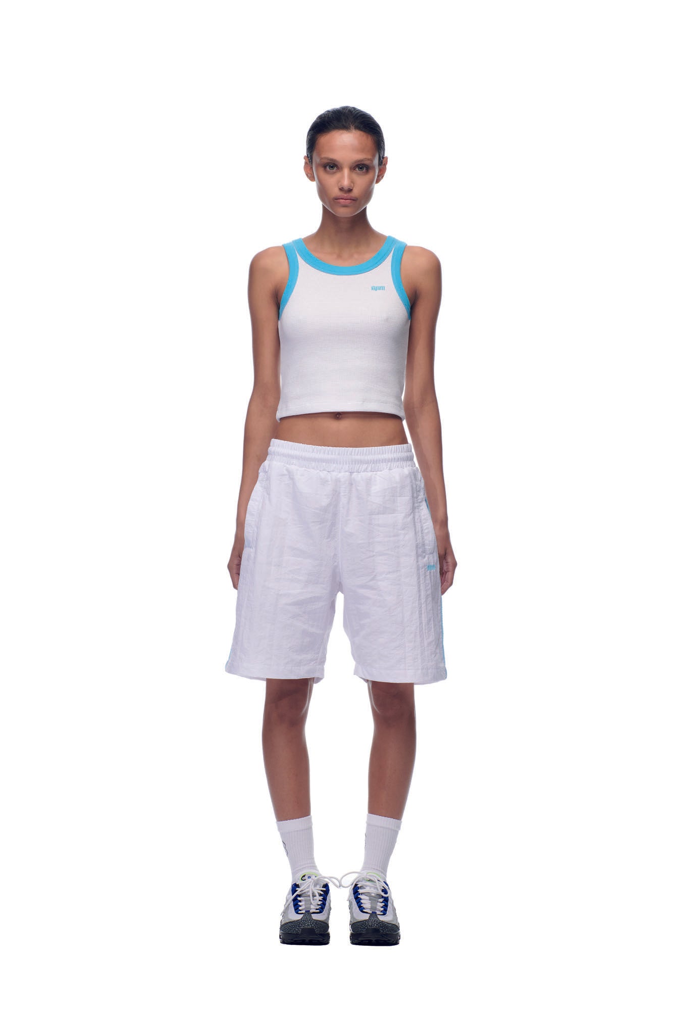 A woman stands facing forward against a white background, wearing 6PM's NH NYLON SHORTS WHITE, a white crop tank with blue trim, white socks, and black-and-white sneakers.