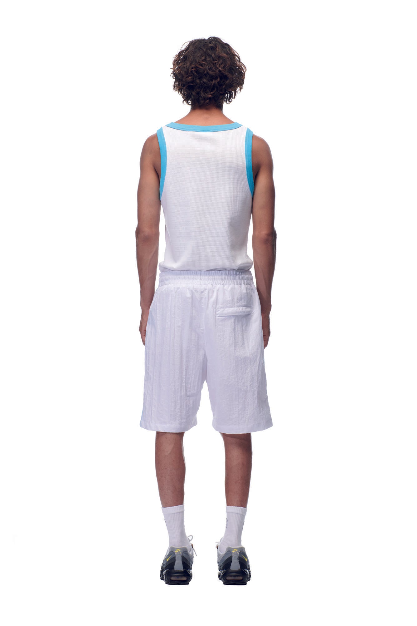 A person with curly hair stands facing away against a white background, wearing 6PM’s NH NYLON SHORTS WHITE, a sleeveless white tank top with blue trim, white socks, and black sneakers.
