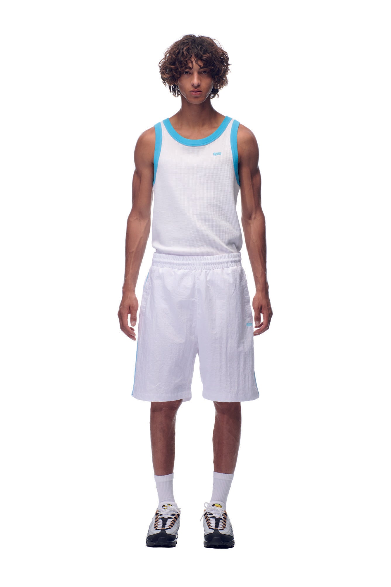 A young man stands facing forward in a white tank top with blue trim, 6PM NH NYLON SHORTS WHITE, white socks, and black-and-white sneakers against a plain white background.