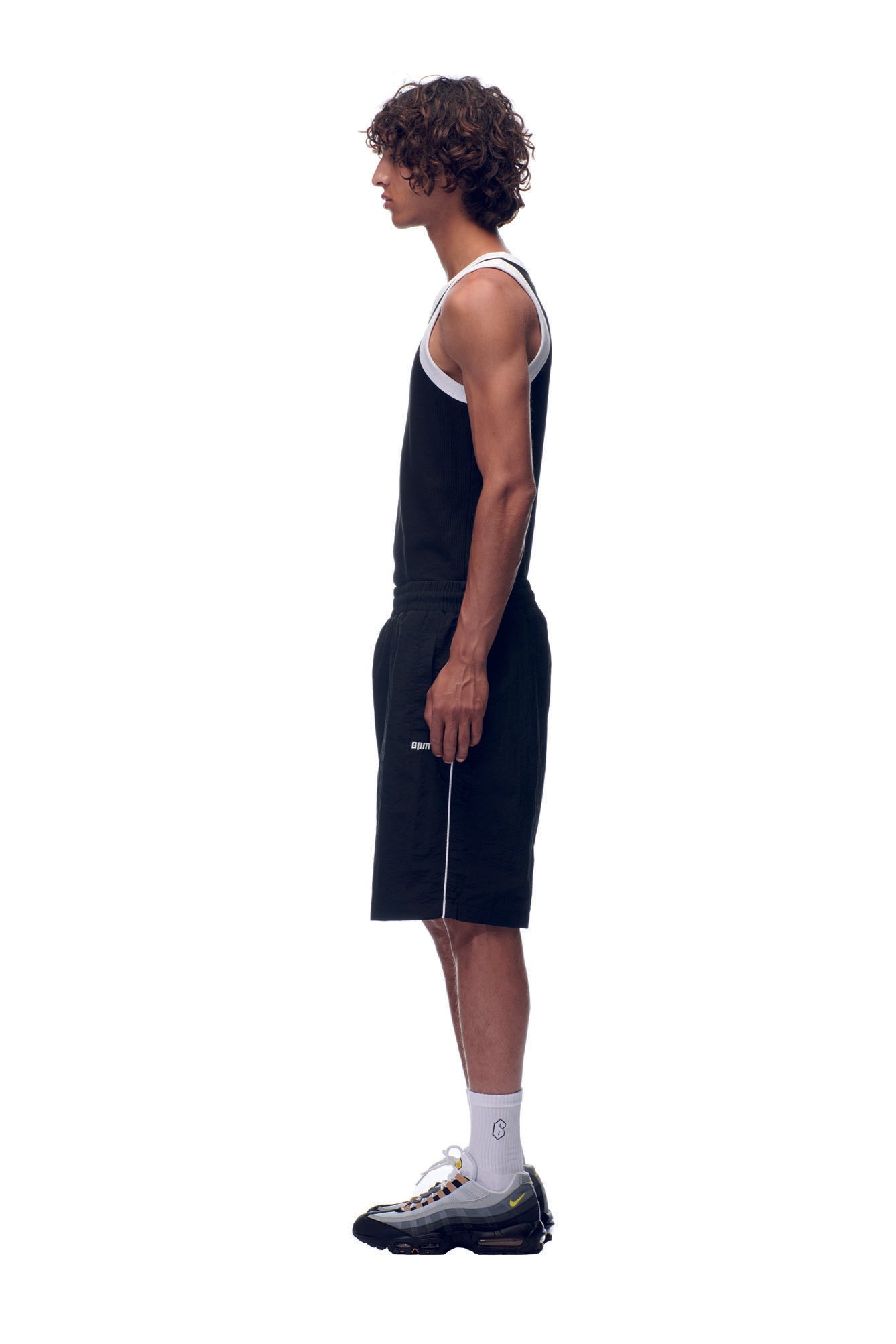 A man with curly hair stands in profile against a white background, wearing a black tank top, 6PM NH NYLON SHORTS BLACK, white socks, and black sneakers.