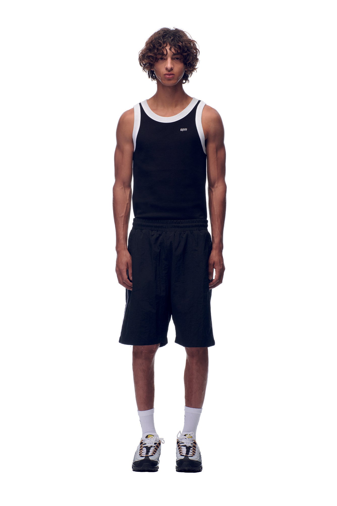 A man with curly hair wears 6PM NH NYLON SHORTS BLACK, a black tank top with white trim, white socks, and multicolored sneakers, standing against a plain white background.