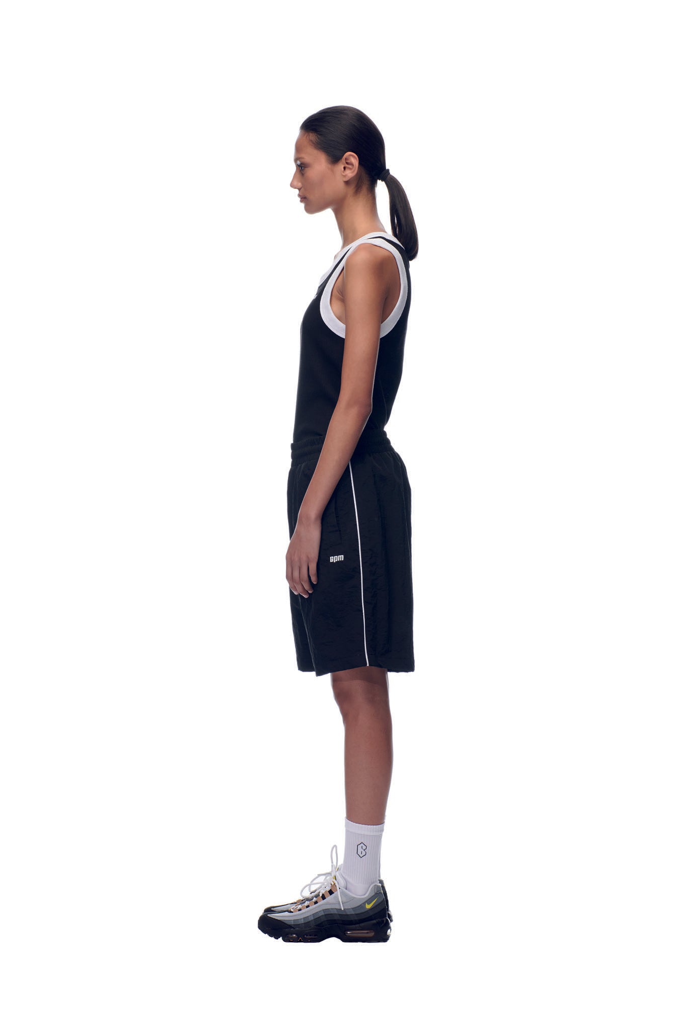 A person with dark hair in a low ponytail stands in profile against a white background, wearing 6PM’s NH NYLON SHORTS BLACK, a black tank top, black sneakers, and white socks.