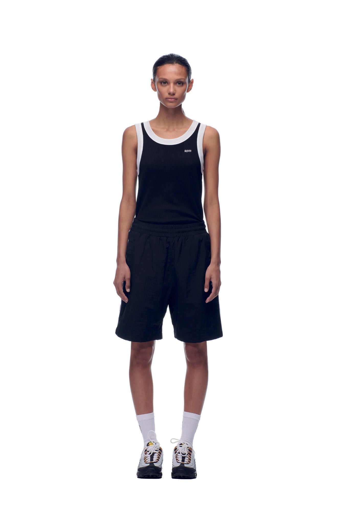 A person stands facing forward against a white background, wearing 6PM NH NYLON SHORTS BLACK, a black tank top with white trim, white crew socks, and black-and-white sneakers.