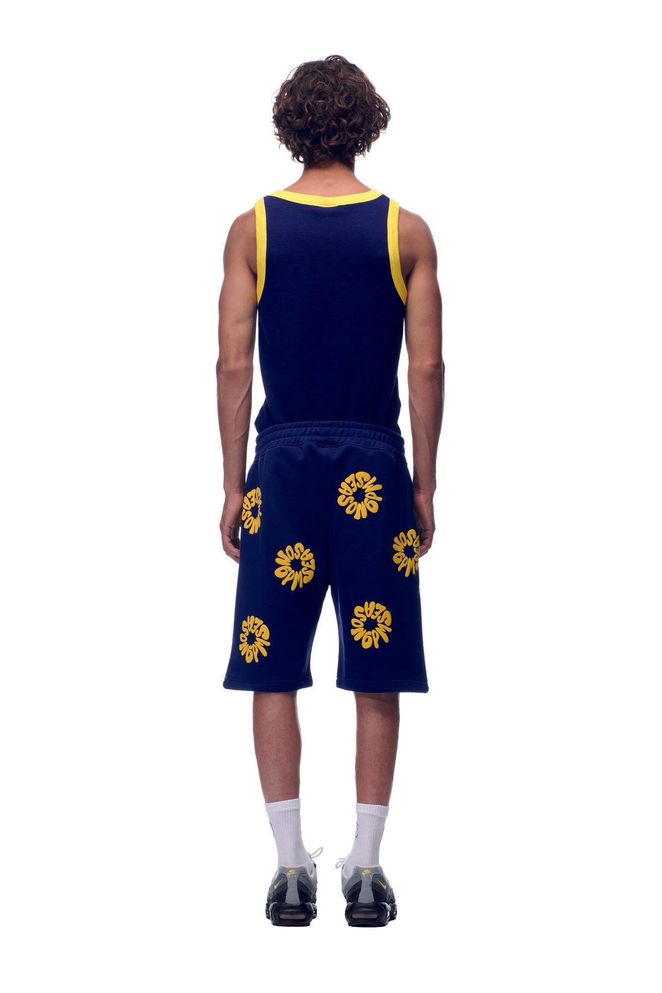 A person faces away, wearing 6PM's FLOWER SHORTS NAVY with yellow floral patterns, paired with a navy tank top with yellow trim, short curly hair, white socks, and black athletic shoes, set against a white background.