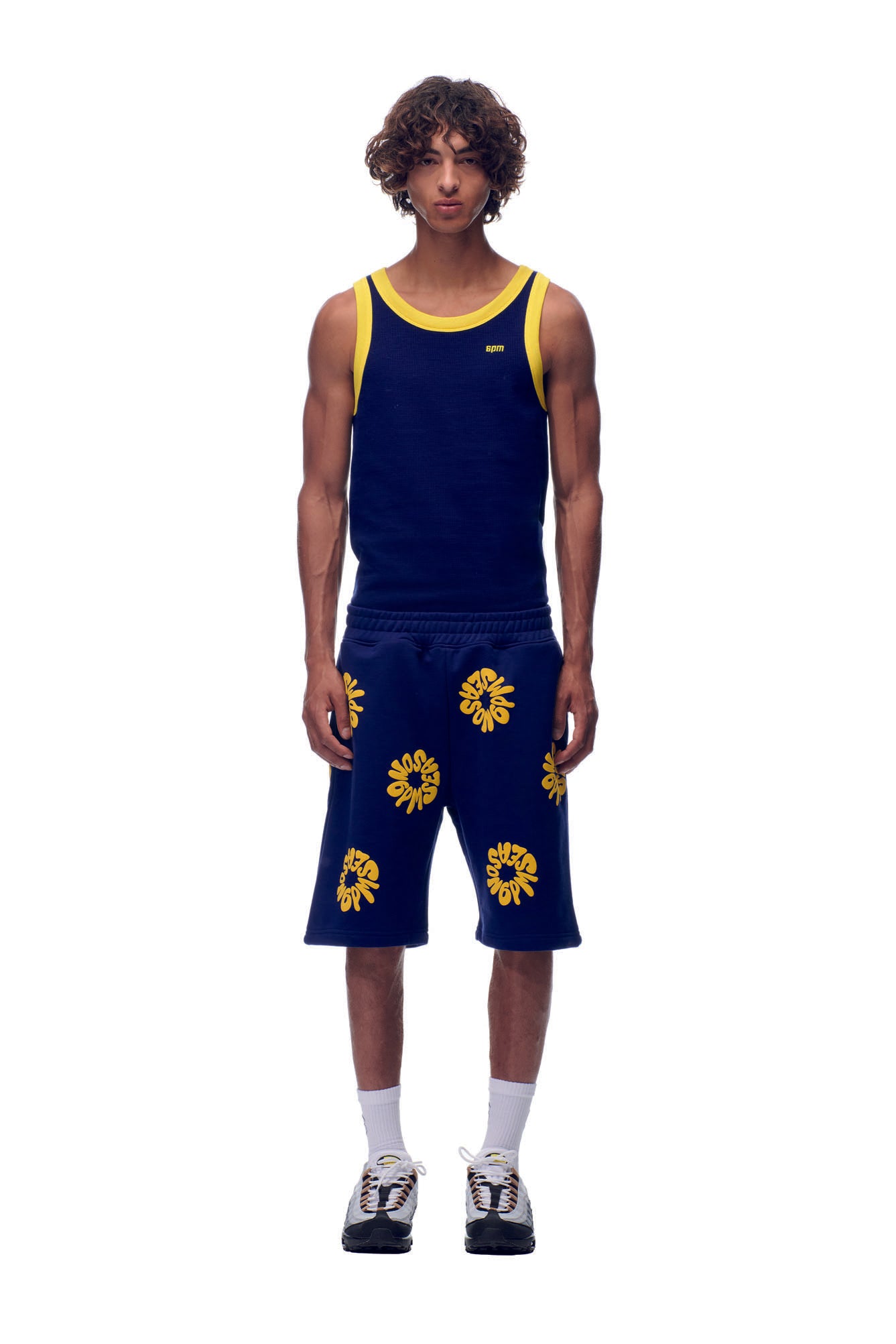 A young man stands against a white background wearing 6PM's FLOWER SHORTS NAVY and a matching navy tank top with yellow trim, paired with white socks and black-and-white sneakers.
