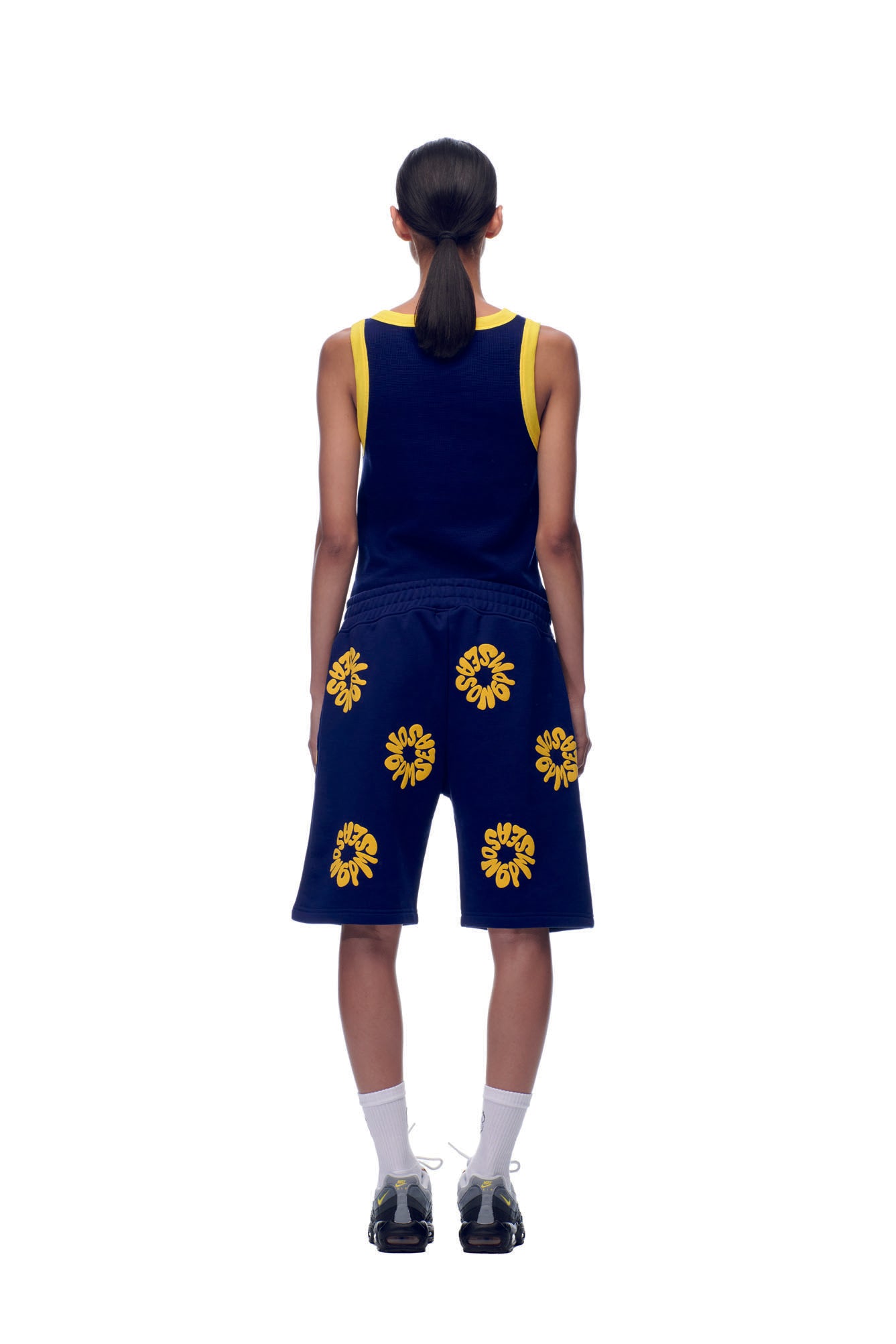 A person faces away, wearing 6PM’s FLOWER SHORTS NAVY with yellow floral patterns, paired with a navy tank top with yellow trim, white socks, black shoes, and their dark hair in a low ponytail.