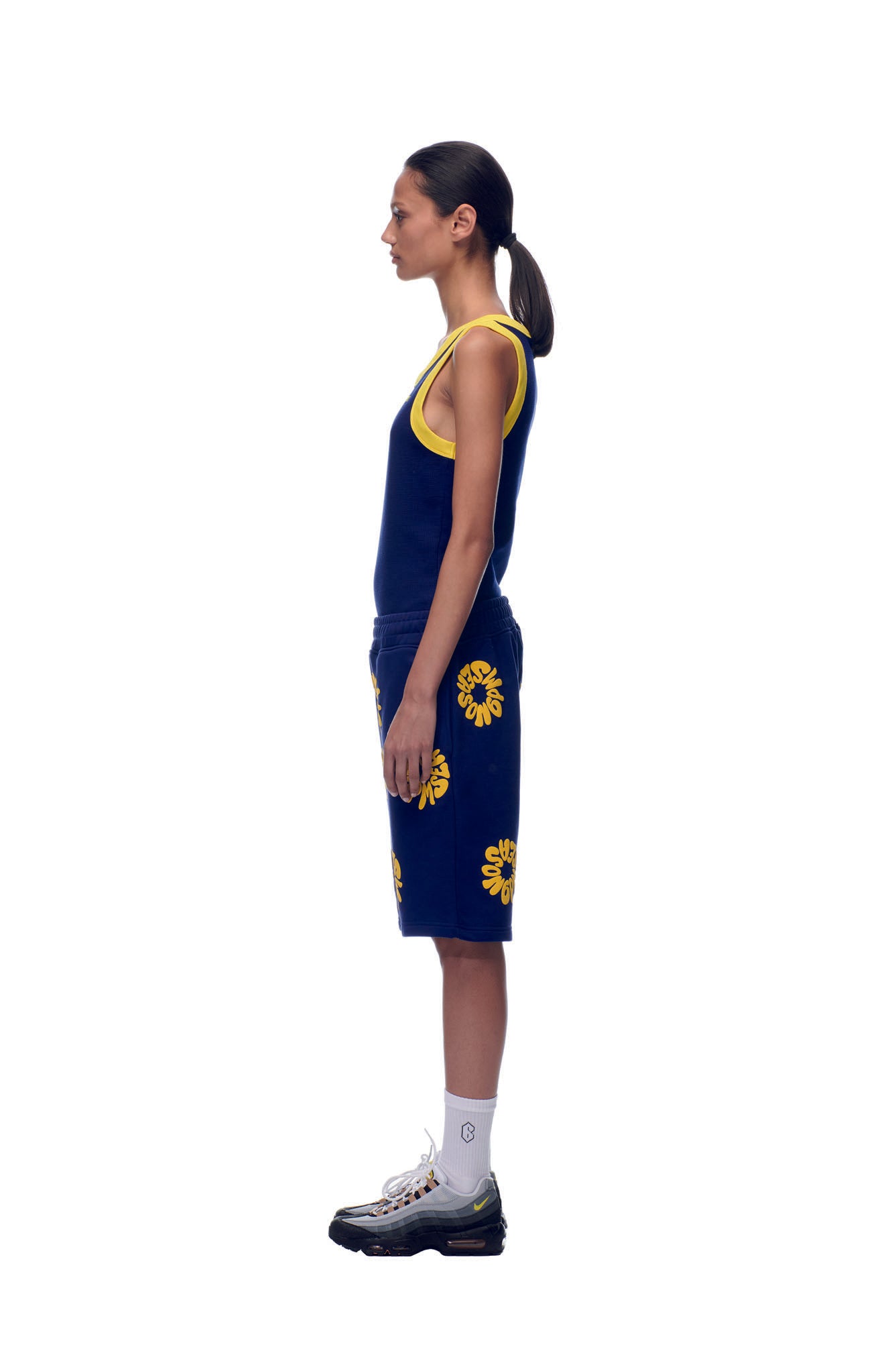 A woman stands in profile against a white background, wearing 6PM’s FLOWER SHORTS NAVY and a matching tank top, paired with white socks and gray sneakers. Her hair is styled in a low ponytail.