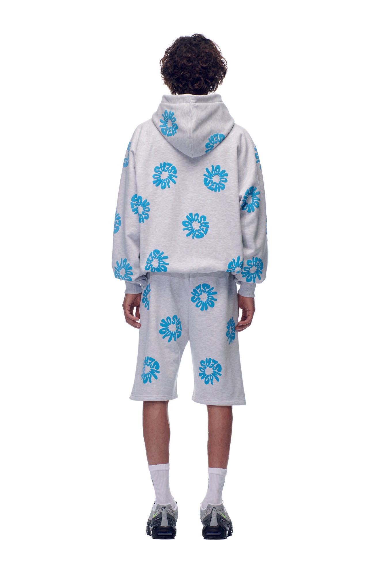 A person with curly hair, seen from behind, wears the 6PM FLOWER HOODIE GREY and matching shorts with blue flower patterns, white socks, and dark sneakers, standing against a white background.