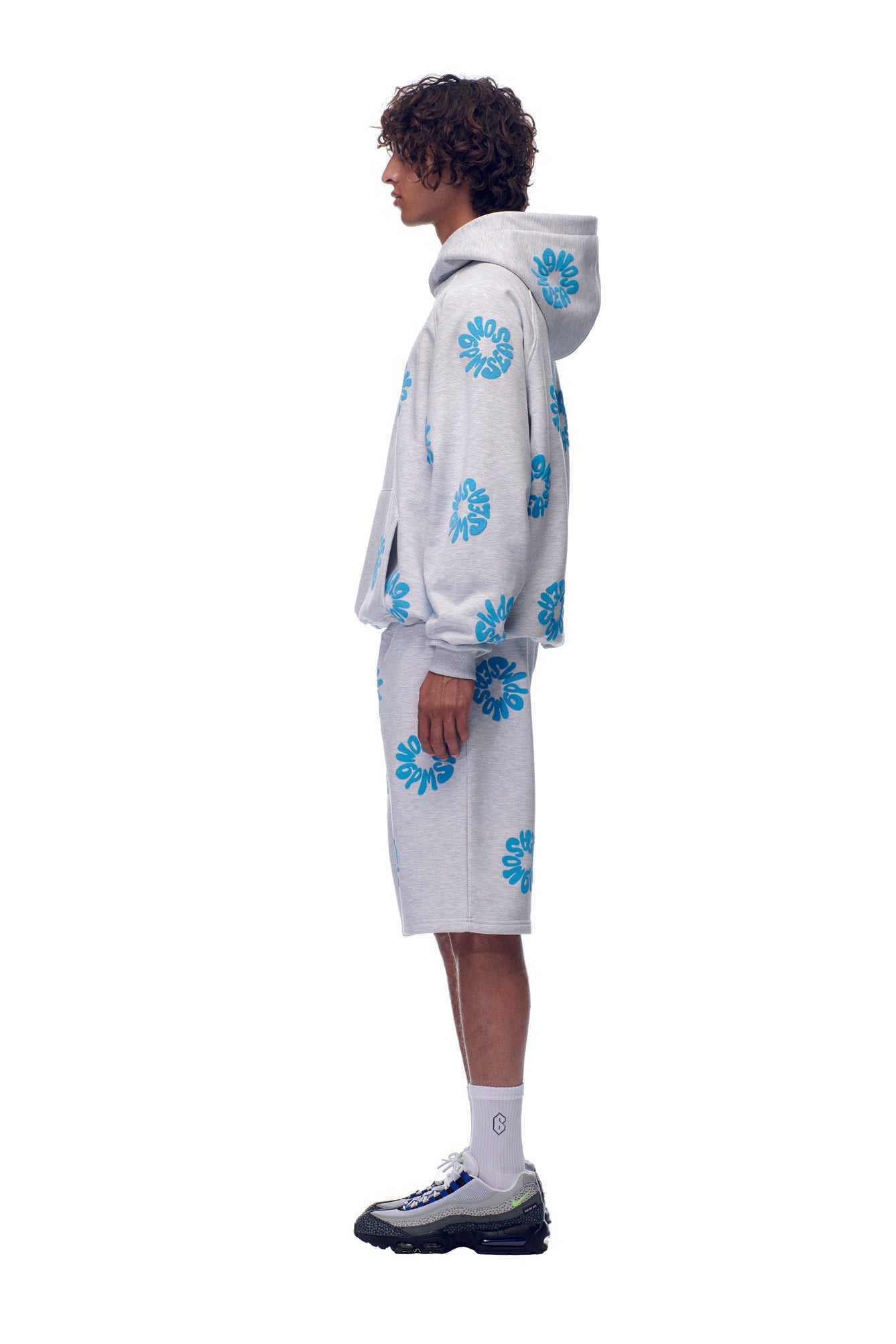 A person stands in profile against a white background, wearing the 6PM FLOWER HOODIE GREY with blue floral shorts, white socks, and black-and-white sneakers.