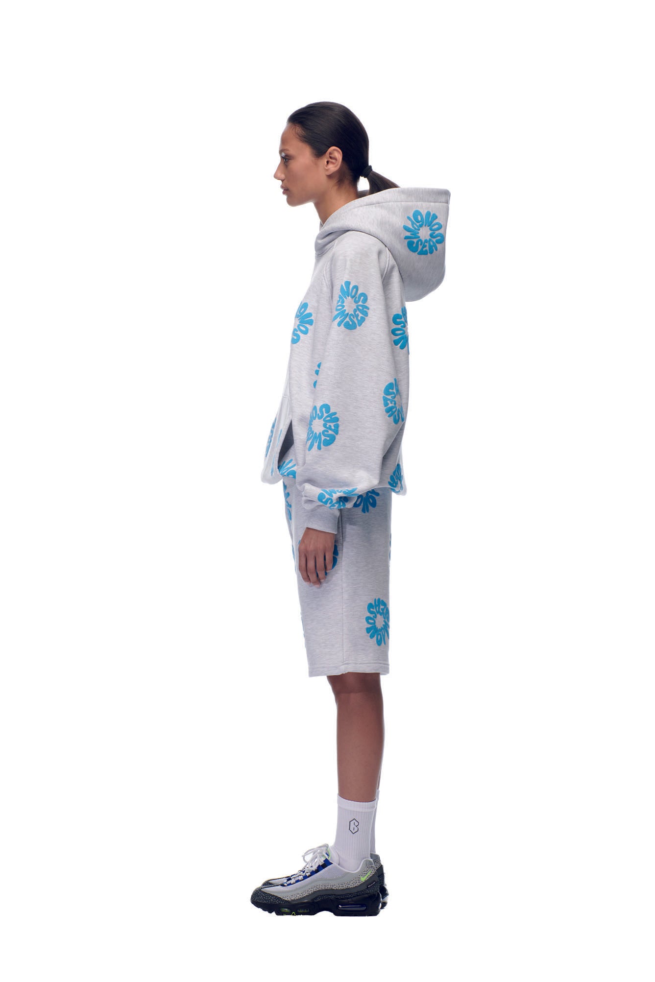 A person stands in profile against a plain white background, wearing the 6PM FLOWER HOODIE GREY with blue floral print shorts, white socks, gray sneakers, and hair tied back in a low ponytail.
