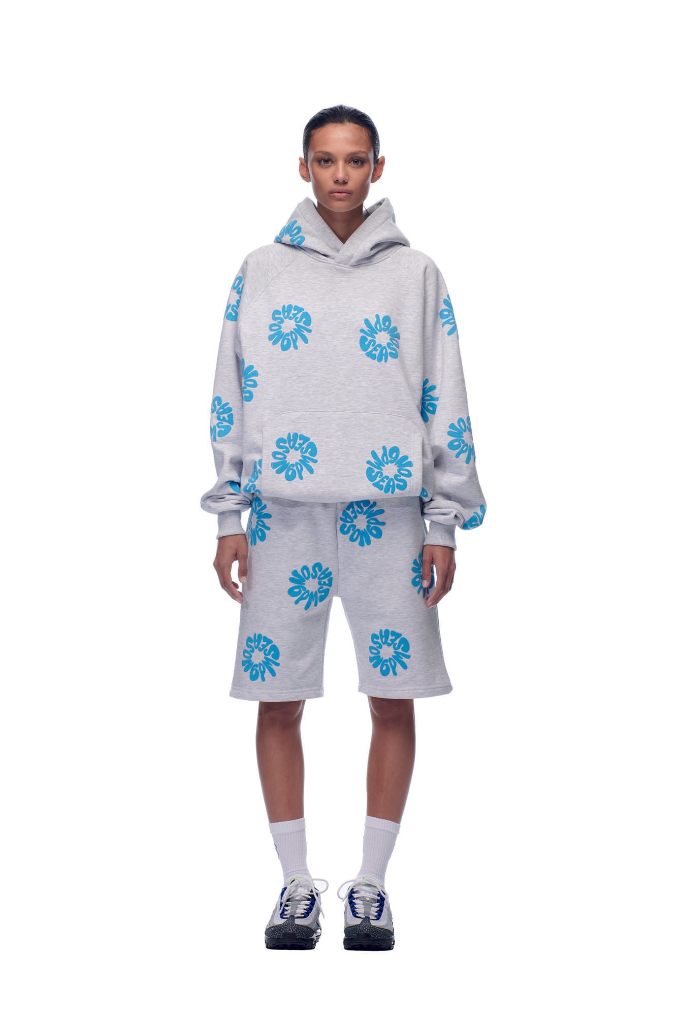 A person faces forward in a 6PM FLOWER HOODIE GREY with matching shorts, both featuring large blue floral prints. They wear white socks and black sneakers, set against a plain white backdrop.