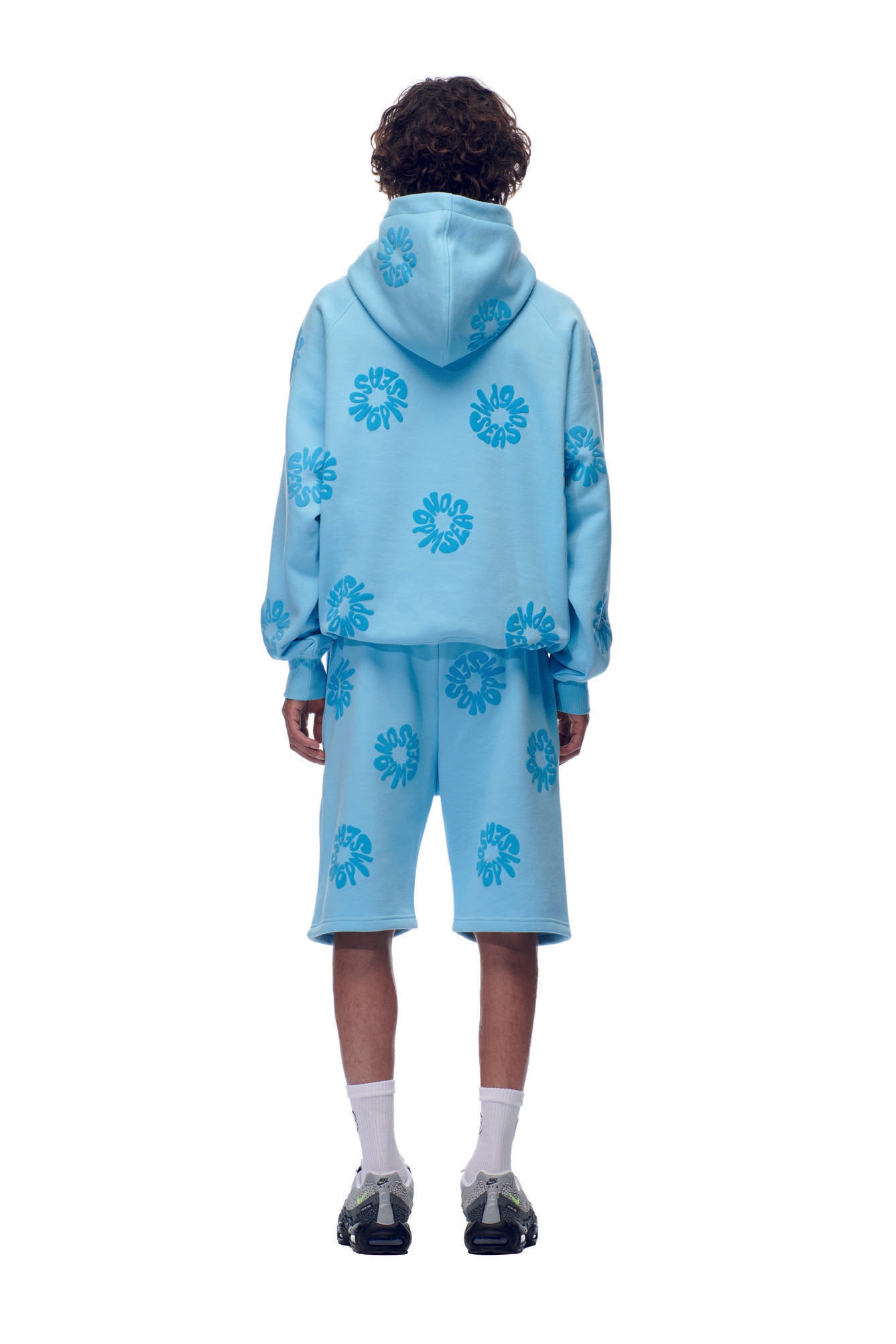 A person with curly hair faces away, wearing the 6PM FLOWER HOODIE BABY BLUE and matching shorts with blue floral patterns, white socks, and black sneakers against a white background.