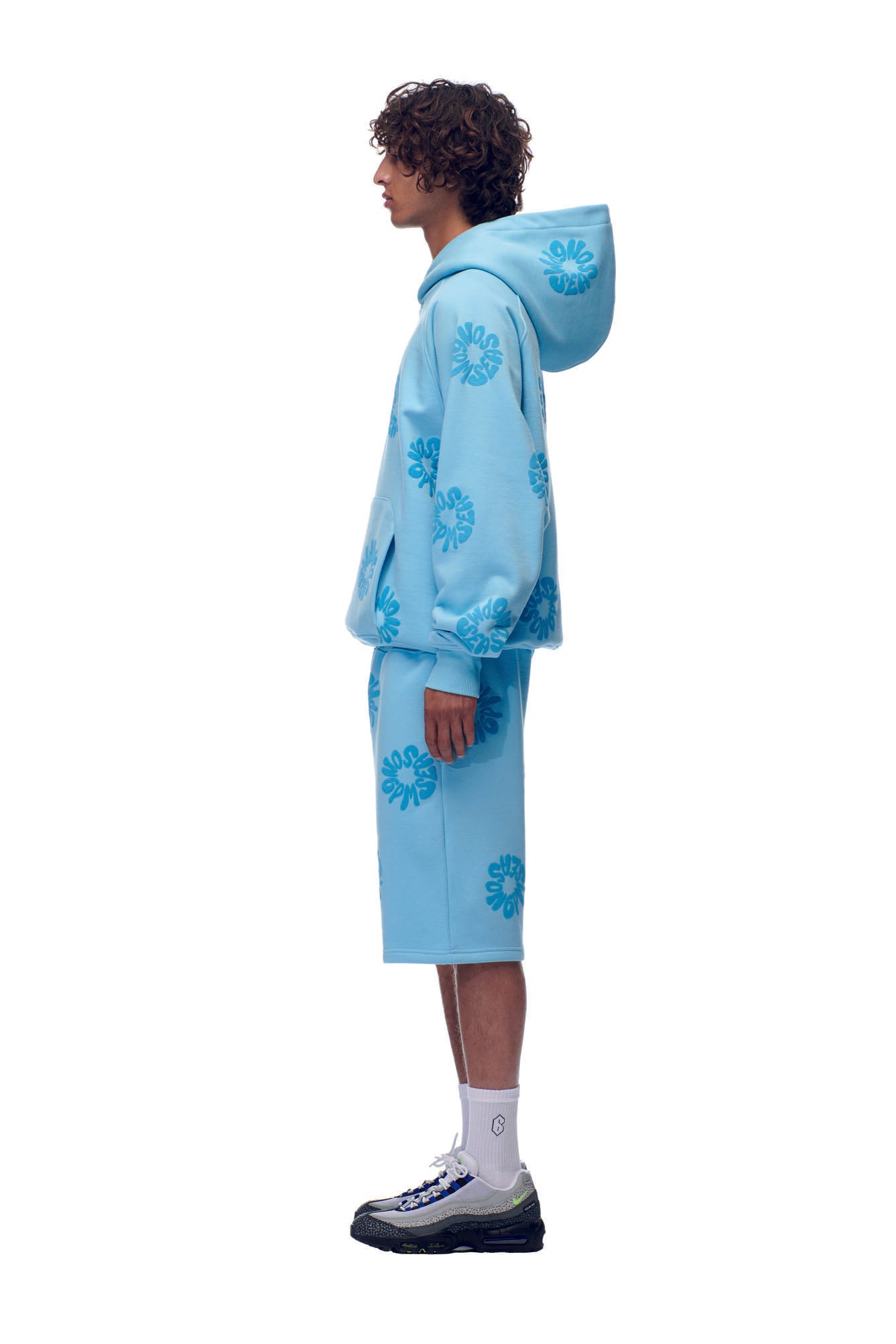 A person in profile wears the 6PM FLOWER HOODIE BABY BLUE with matching floral shorts, white socks, and black sneakers against a plain white background.