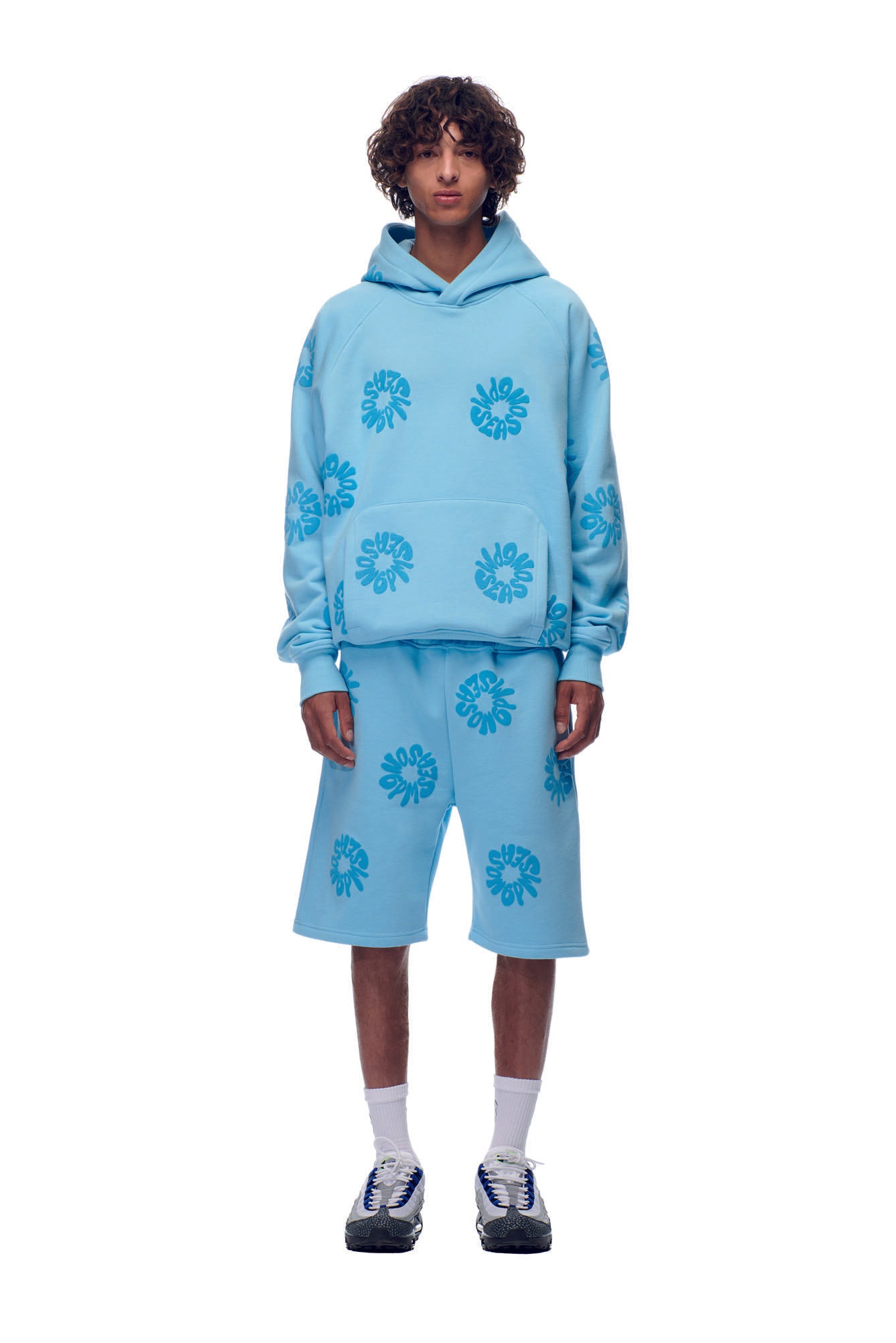 A person wearing the 6PM FLOWER HOODIE BABY BLUE with matching knee-length shorts featuring blue abstract floral patterns, paired with white socks and black sneakers, stands against a plain white background.