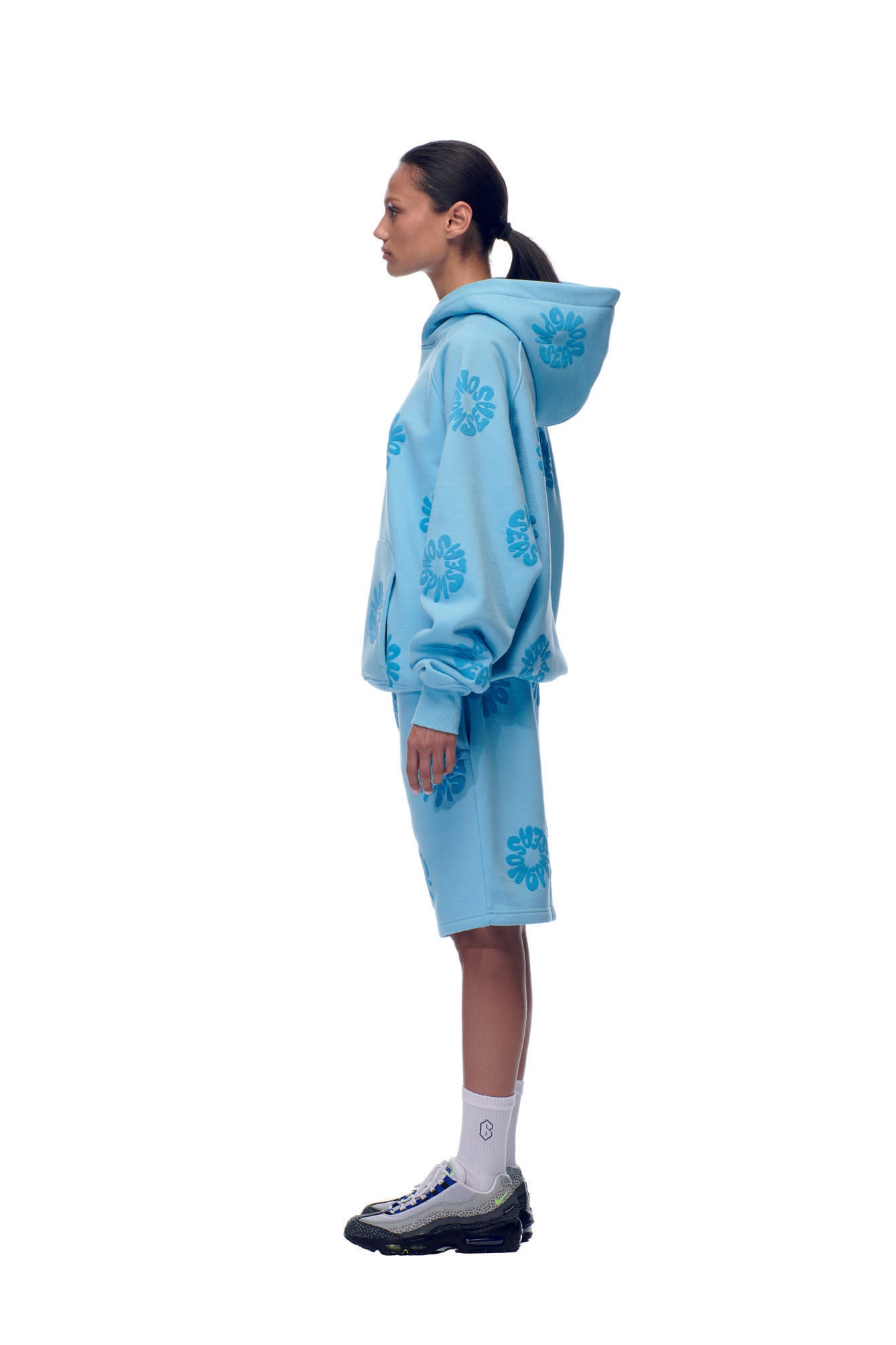 A woman stands in profile against a white background, wearing the 6PM FLOWER HOODIE BABY BLUE set—featuring a floral hoodie and matching shorts—paired with white socks and black and white sneakers.